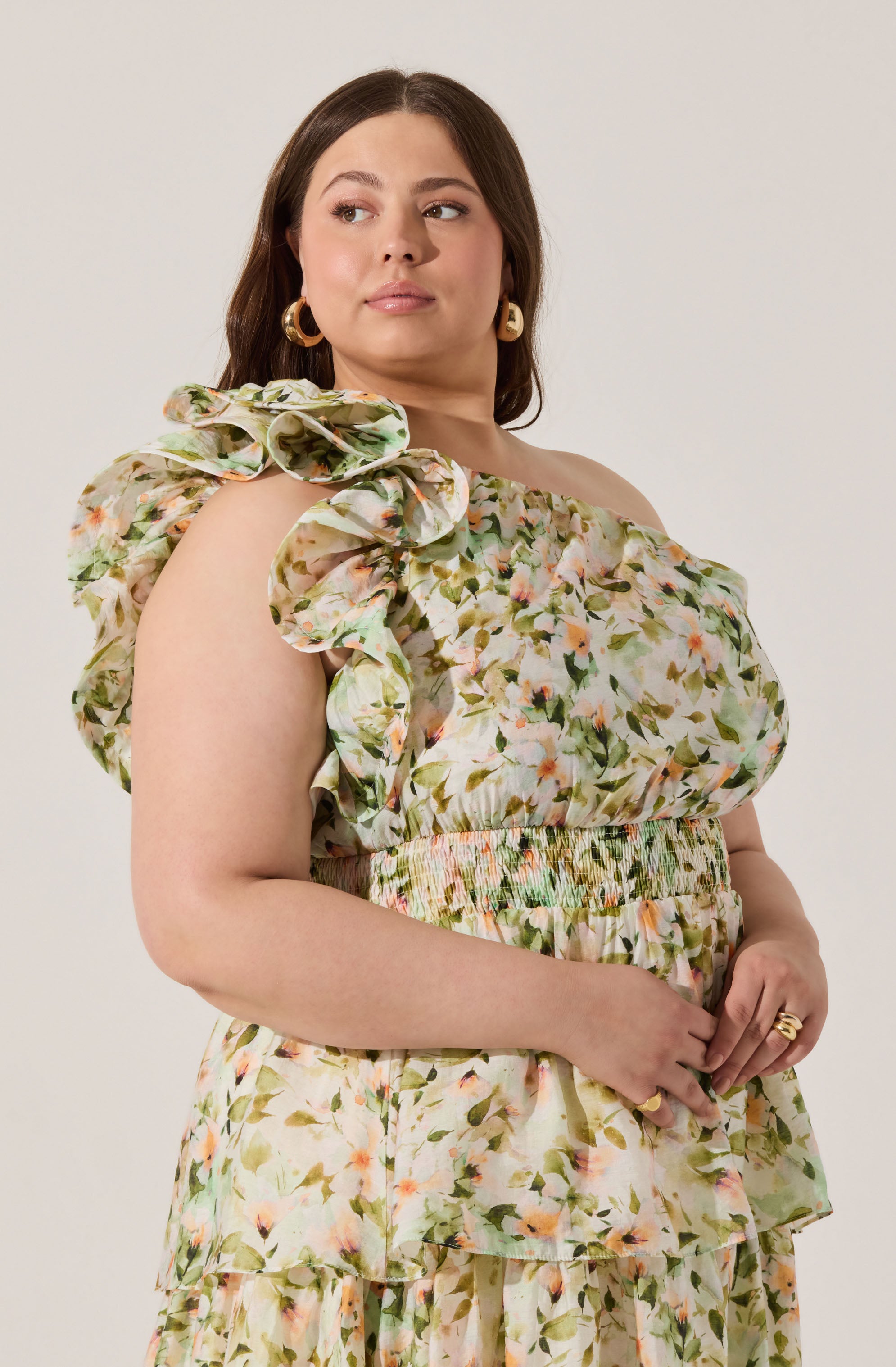 A woman is wearing a floral print dress with an off-the-shoulder design. The dress features ruffled sleeves and a smocked waist. The fabric has a light background with a pattern of green leaves and pink, orange, and yellow flowers. She is also wearing large, gold hoop earrings and a gold ring.