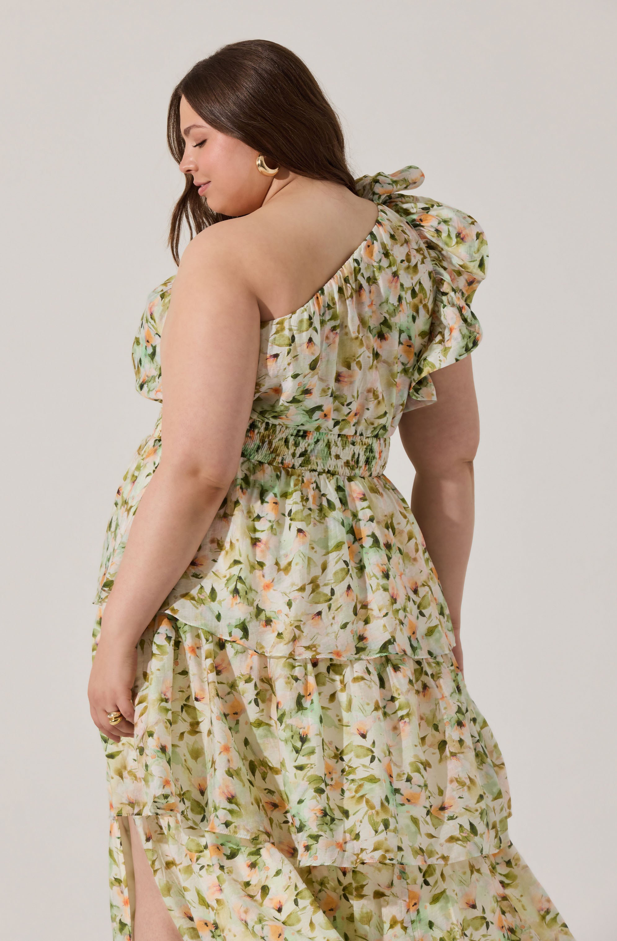 A woman is wearing a sleeveless, floral-patterned dress with ruffled details on the shoulder and a fitted, smocked waist. The fabric features a light, pastel-colored print with various shades of green, peach, and pink.