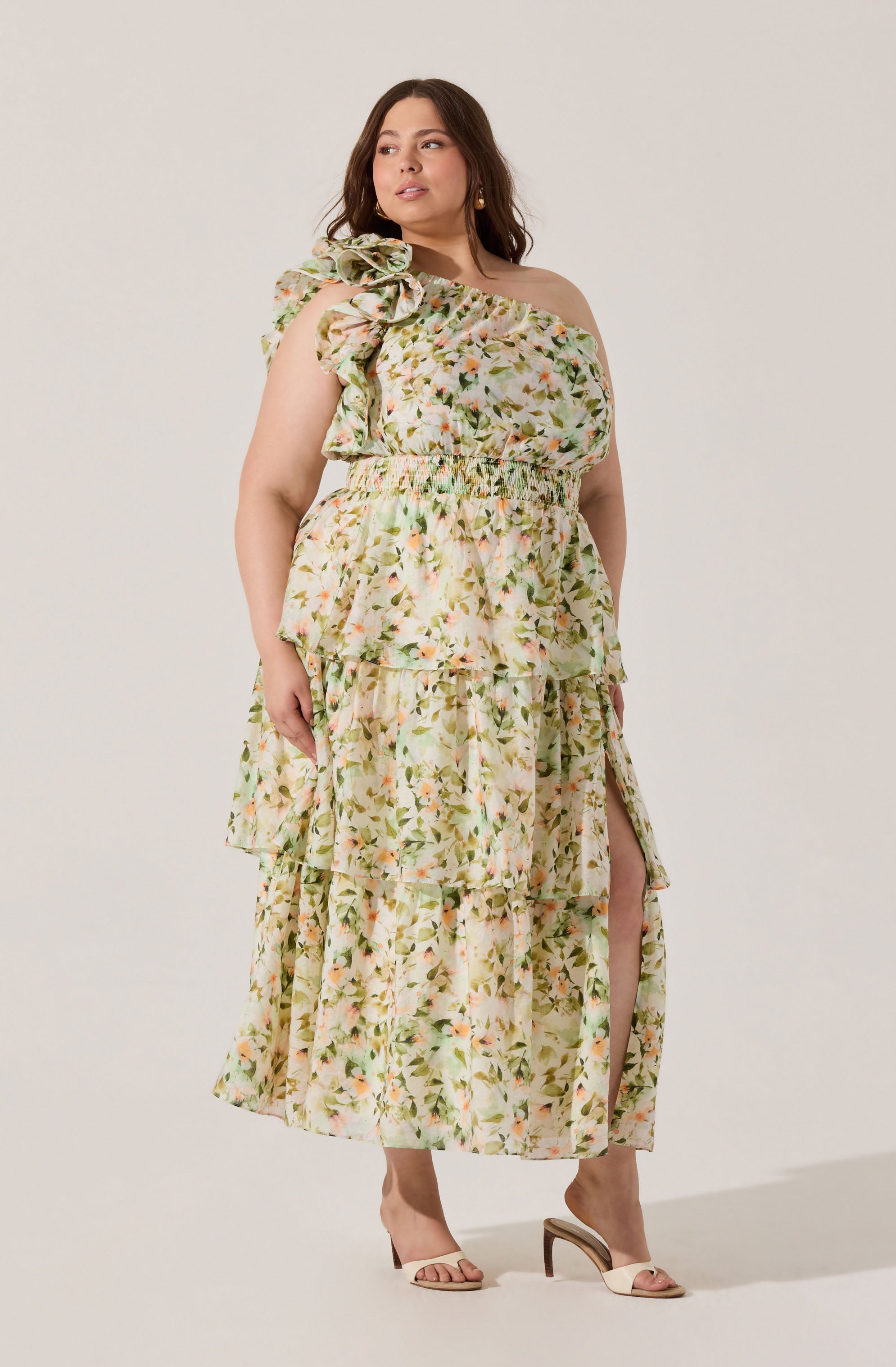 A woman is wearing a floral strapless dress with a ruffled neckline and an asymmetrical, tiered skirt that features a slit on one side. The dress has a light, pastel color palette with a pattern of small flowers and leaves. She is also wearing open-toe high heels with a thin ankle strap.