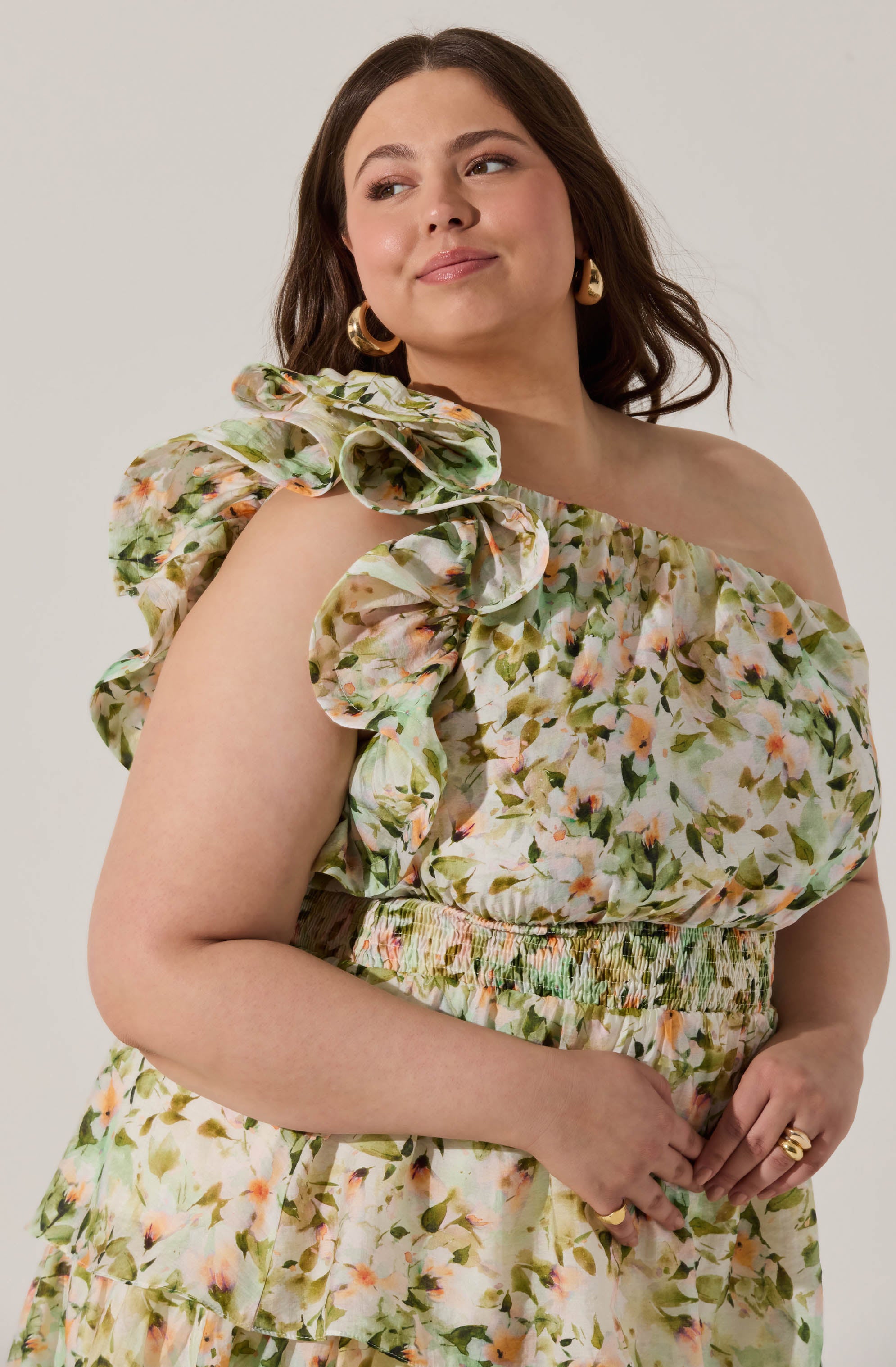 A woman is wearing a floral dress with a ruffled, puffed sleeve on her left shoulder. The dress features a multicolored pattern of green, peach, and pink flowers and has an elastic waistband. She is accessorized with gold hoop earrings and a gold ring.
