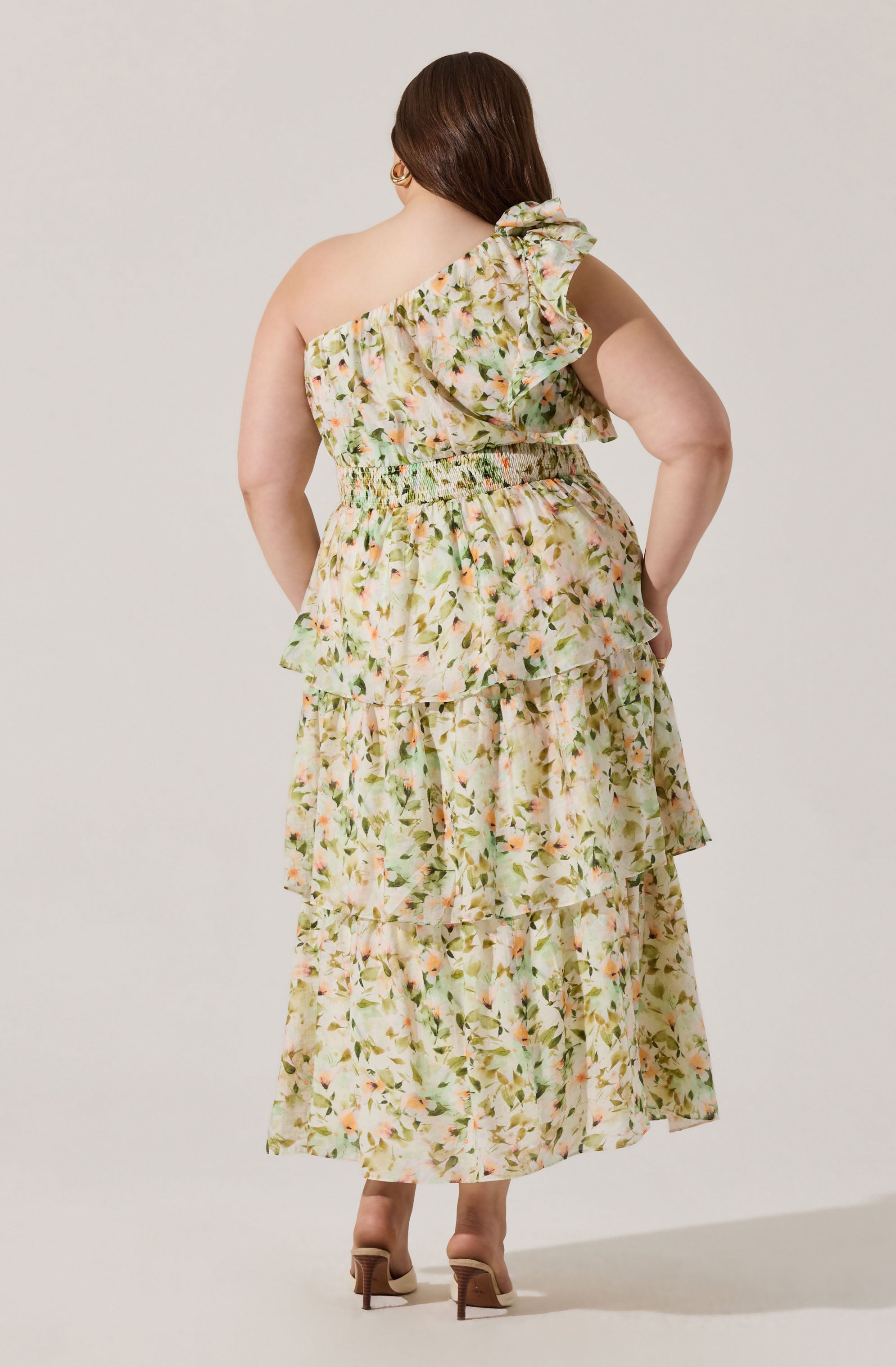 A woman is wearing a long, flowy dress with a floral print in soft pastel colors. The dress features a one-shoulder design with a large ruffle detail on the shoulder and around the neckline. The waist is gathered with elastic, creating a cinched effect, and the skirt is tiered with multiple ruffled layers, giving it a layered, voluminous look. The fabric has a light, airy appearance suitable for warm weather. She is also wearing beige high-heeled shoes.