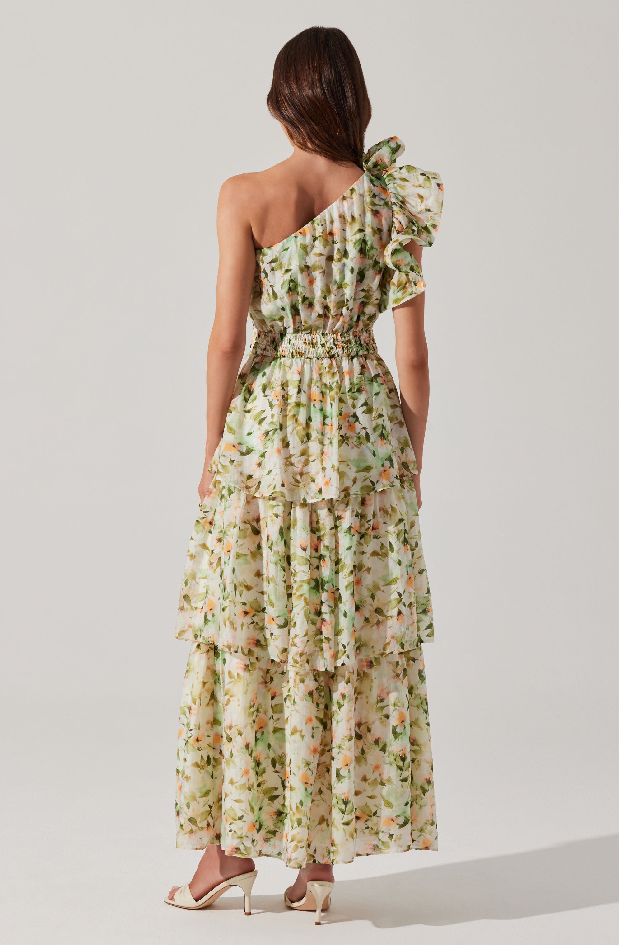A woman is wearing a long, flowing floral dress with a one-shoulder design. The dress features a ruffled strap on the left shoulder and an elastic waistband. The fabric has a light, airy appearance with a pattern of soft-colored flowers and green leaves throughout.