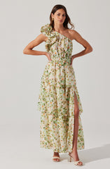 A woman is wearing a long, flowy dress with a floral pattern in shades of green, beige, and pink. The dress features an asymmetrical, one-shoulder design with a large ruffle on the shoulder. It has an elastic waistband and a high slit on the left side that reveals her leg.