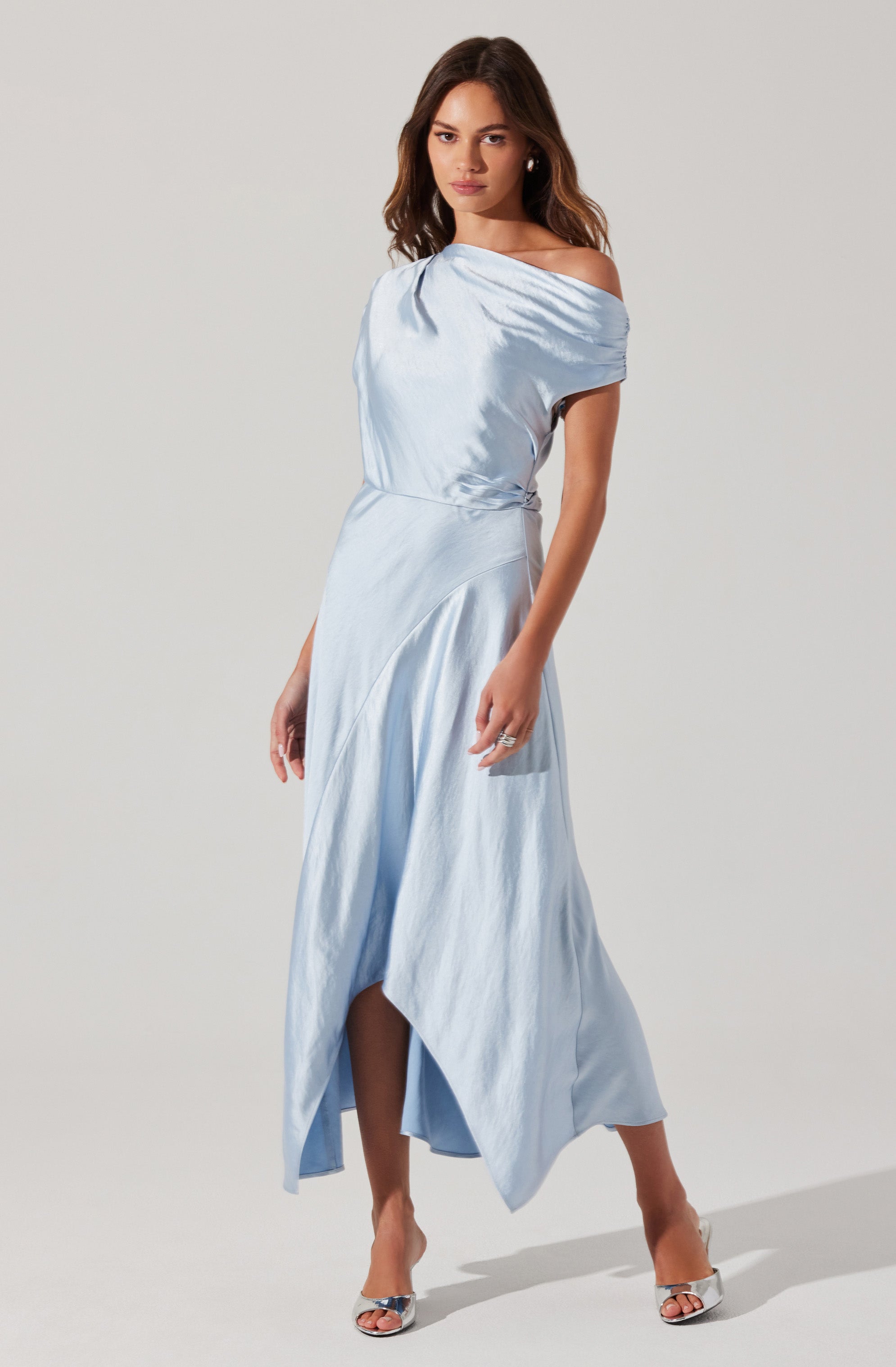 A woman is wearing a light blue satin dress with a draped, off-the-shoulder neckline. The dress has a fitted waist and a flowing, asymmetrical hemline that is longer in the back and shorter in the front. She is also wearing open-toe high heels with a metallic finish and has earrings and rings as accessories.