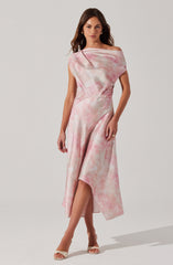 A woman is wearing a sleeveless, off-the-shoulder dress with a pink and white tie-dye pattern. The dress has a fitted waist and a flowing, asymmetrical hemline that is longer in the back and higher in the front, revealing her legs. She is also wearing open-toe heels and gold hoop earrings.