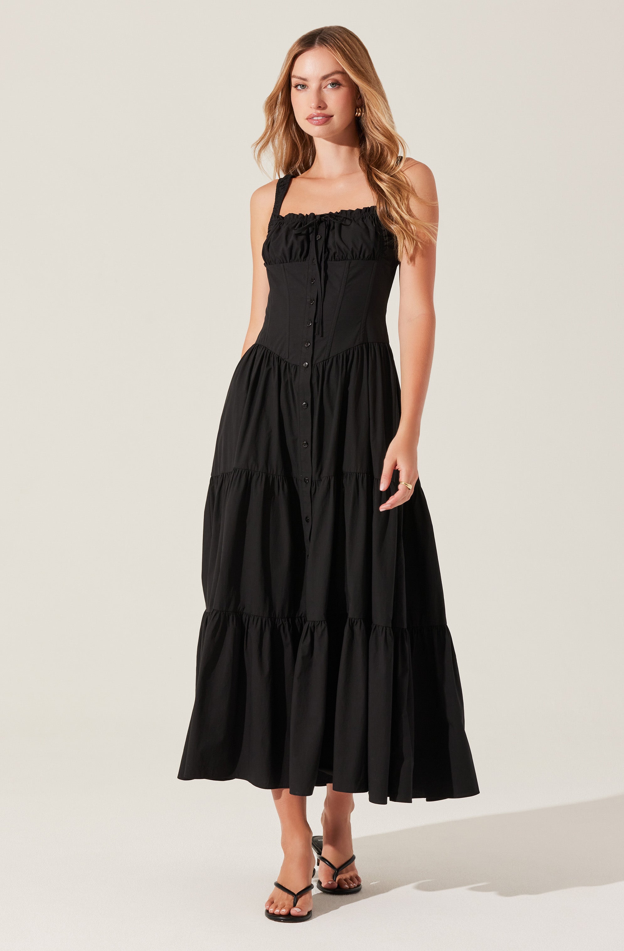 A woman is wearing a black sleeveless, floor-length dress with a fitted bodice that features a ruffled neckline and button-down front. The dress has a tiered skirt with gathered fabric at multiple levels, creating a flowing silhouette. She is also wearing black strappy sandals and small hoop earrings.