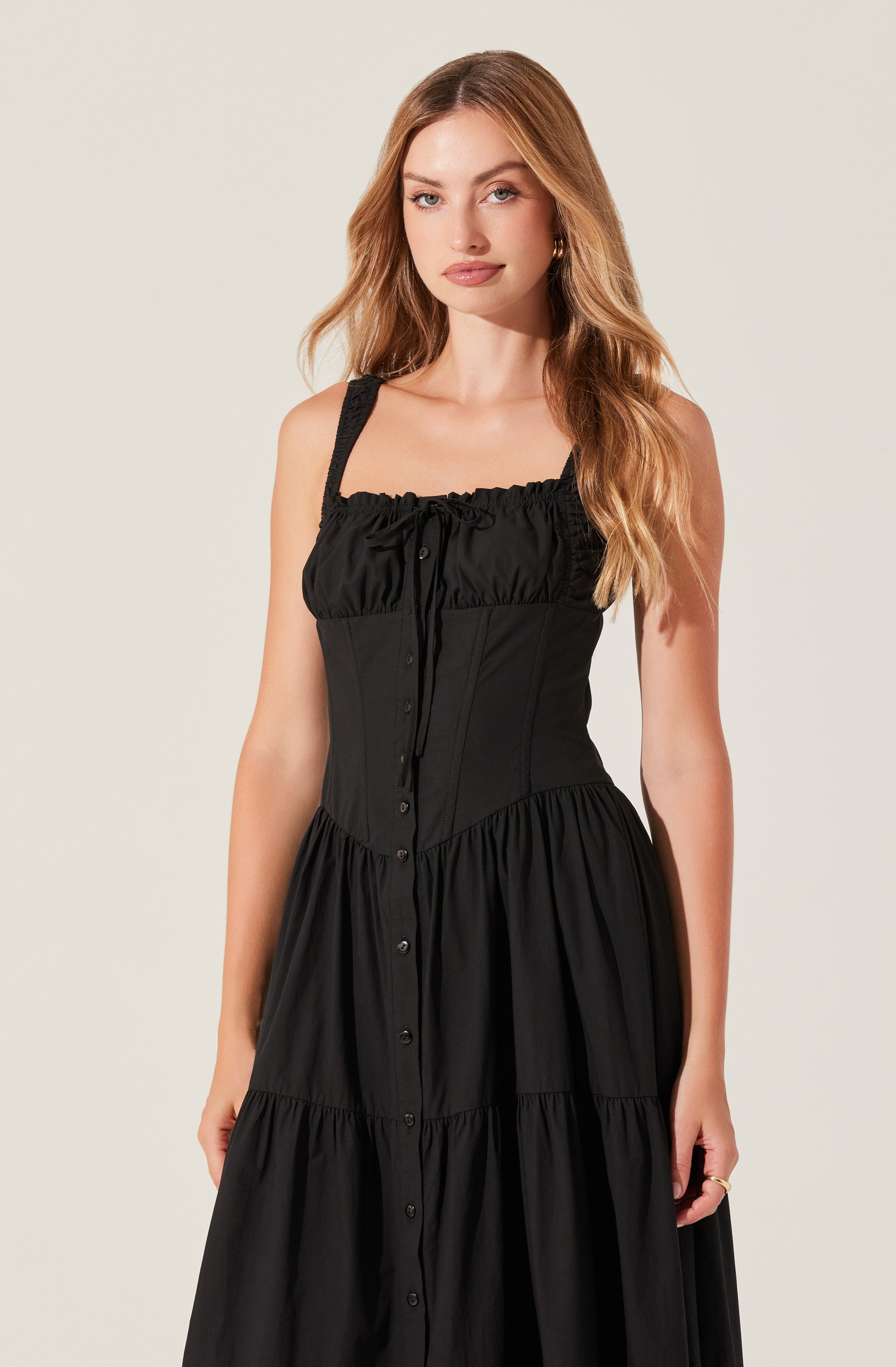 A woman is wearing a sleeveless black dress with wide shoulder straps. The dress features a gathered, ruffled neckline and a fitted bodice with vertical seams. The center front has a row of buttons running from the neckline to the hem. The skirt of the dress is full and gathered, creating a flowing silhouette.