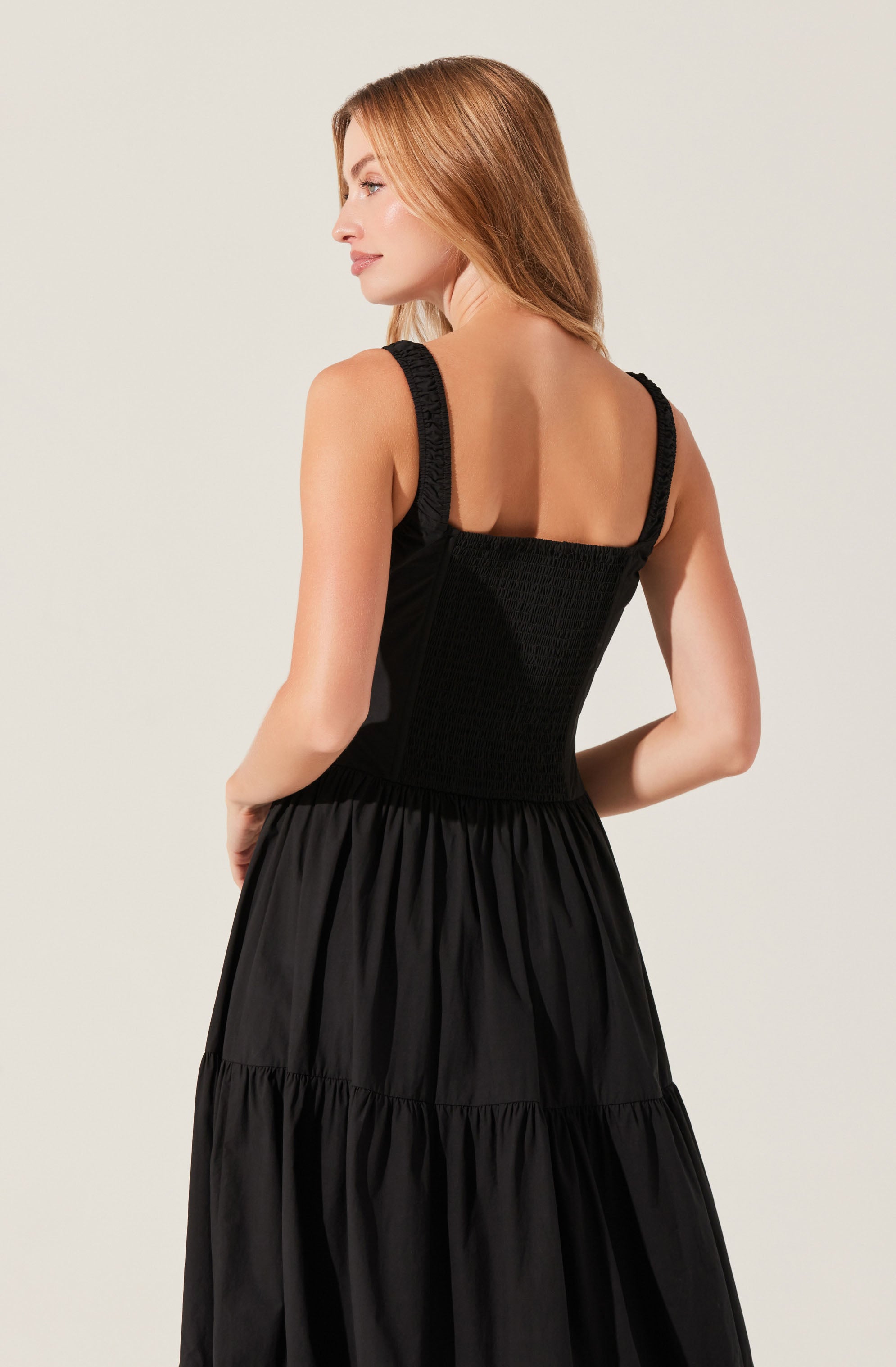 A woman is wearing a black sleeveless dress with wide shoulder straps. The bodice of the dress appears to be fitted and made of a textured fabric, while the skirt is made of a lightweight, gathered material that creates a flowing silhouette.