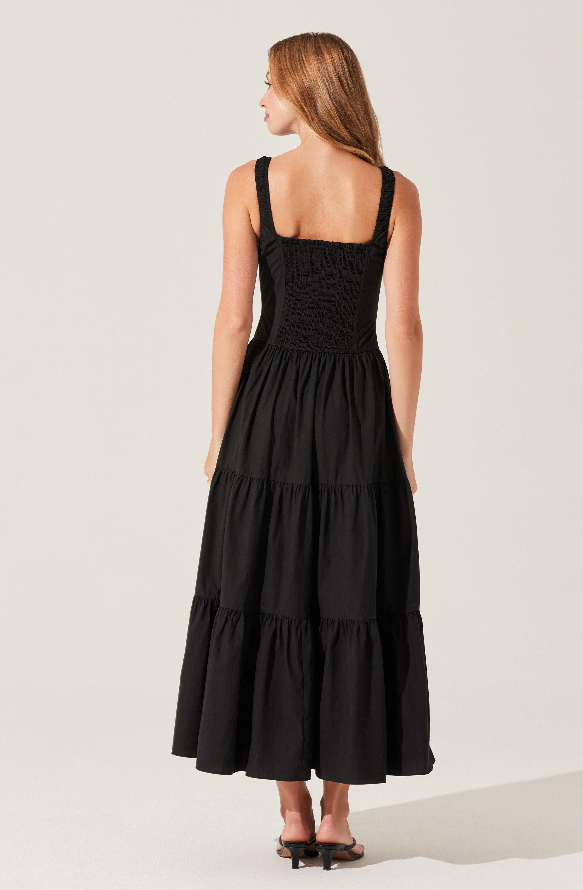A woman is wearing a black sleeveless dress with wide shoulder straps. The top part of the dress features a textured, smocked fabric, while the skirt is long and gathered into multiple tiers that reach down to her ankles. She is also wearing black high-heeled sandals.