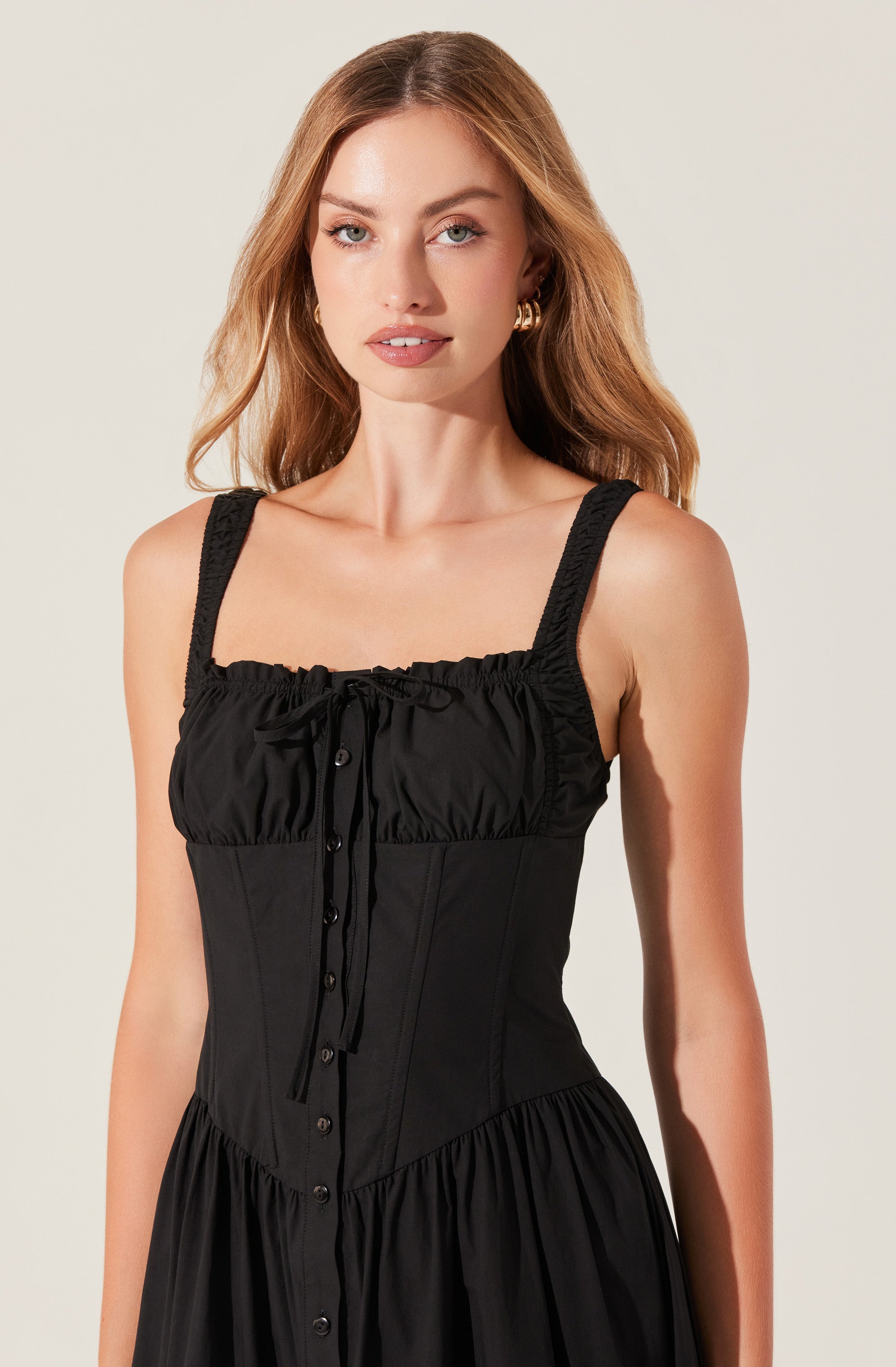 A woman is wearing a black sleeveless dress with wide, textured straps. The bodice features gathered fabric at the top with a drawstring detail and buttons down the front. The skirt part of the dress is gathered and flowing, creating a relaxed look.