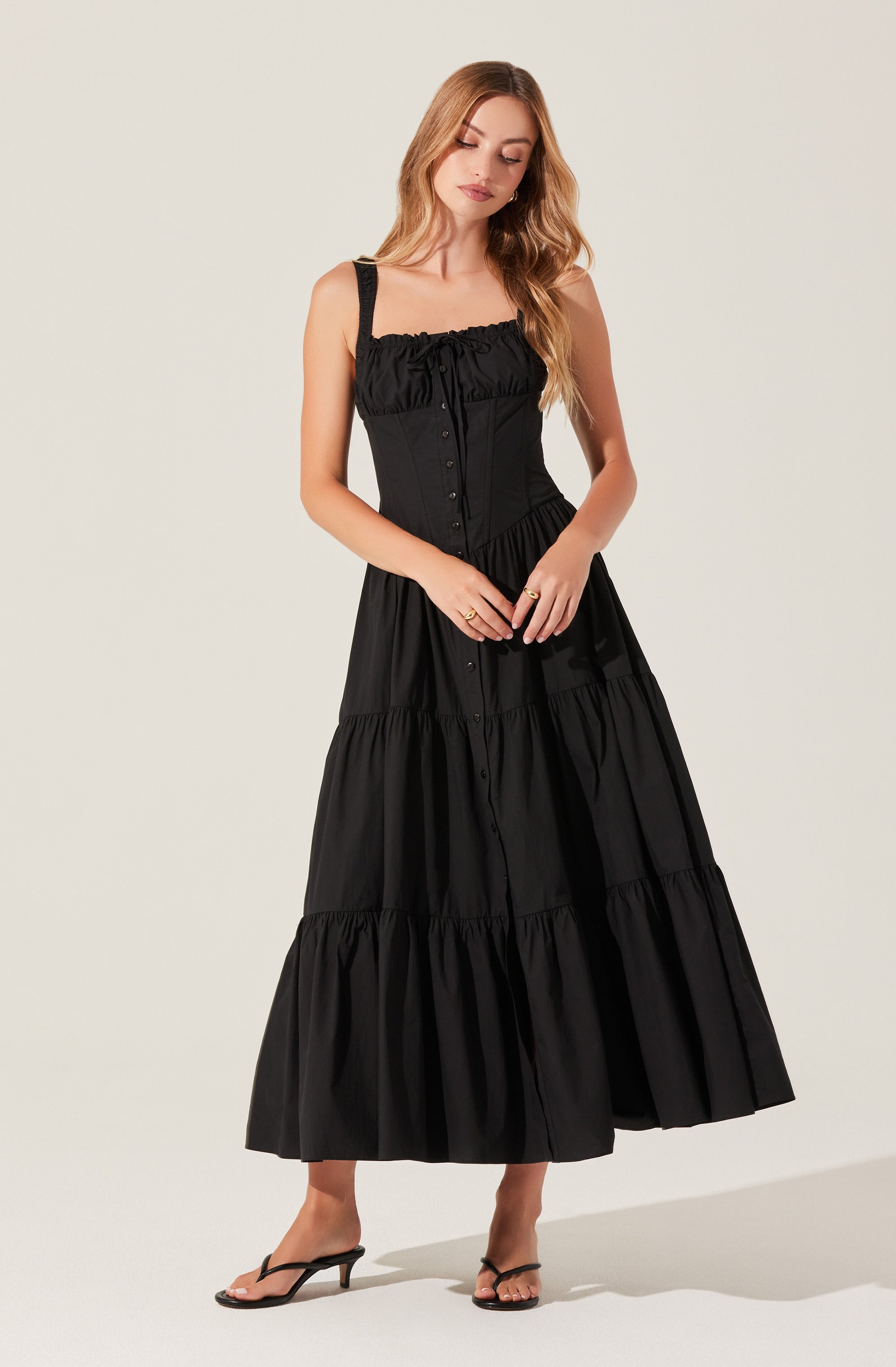 A woman is wearing a long, black, sleeveless dress with a ruffled, square neckline. The dress features a fitted bodice with button details down the front and a tiered, flowy skirt. She completes the look with black high-heeled sandals.