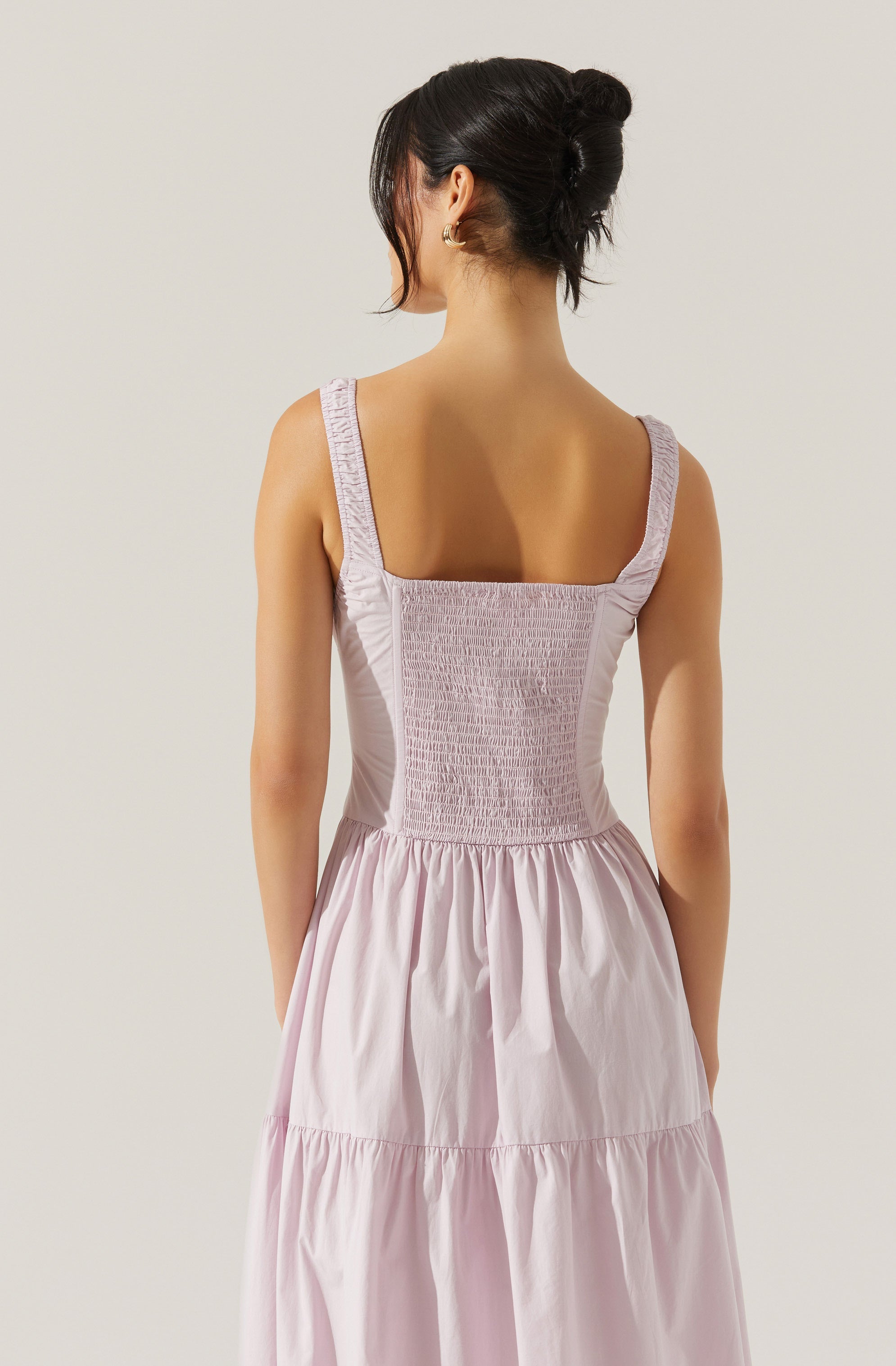 A woman is wearing a sleeveless, light pink dress with wide straps. The top part of the dress features a shirred, textured fabric, while the skirt is gathered and flows loosely with a tiered design. She has dark hair styled in an updo and is wearing small hoop earrings.
