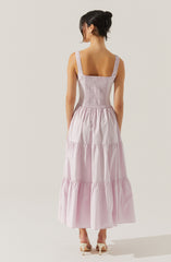 A woman is wearing a sleeveless, light pink maxi dress with wide shoulder straps. The bodice is fitted with gathered or smocked detail, and the skirt is flowing with multiple tiered sections. She is also wearing light-colored high-heeled shoes.