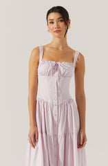 A woman is wearing a sleeveless, light pink sundress with wide straps that have lace details. The dress features a fitted bodice with gathered fabric at the top, a row of buttons running down the front, and a gathered, tiered skirt.
