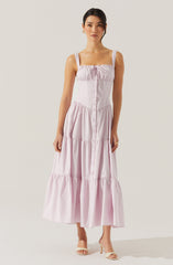 A woman is wearing a sleeveless, light pink, tiered maxi dress with wide shoulder straps and a front button-down design. The dress has a gathered bodice with a small bow at the center of the neckline. She is also wearing open-toed, high-heeled sandals.