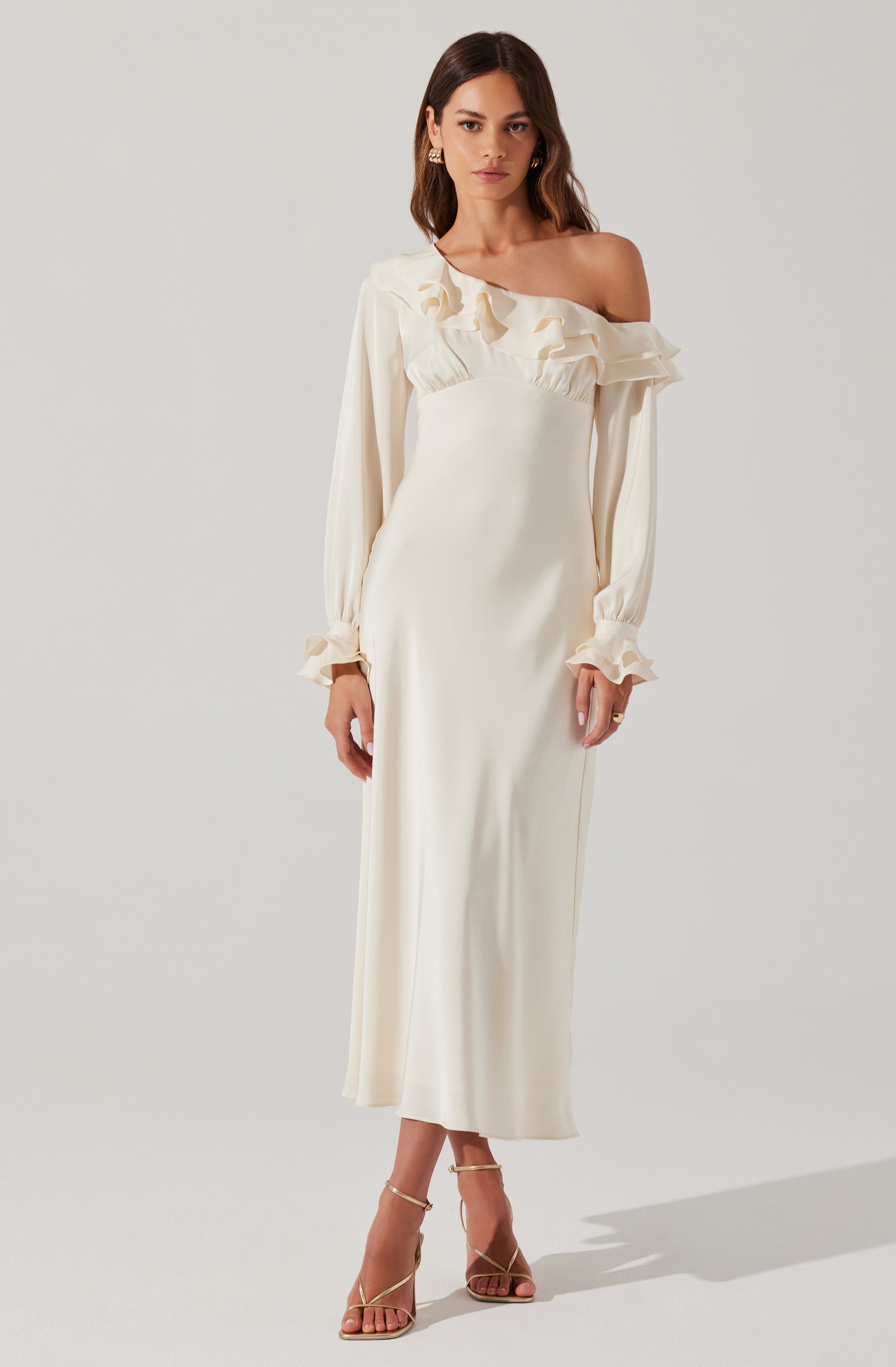 A woman is wearing an elegant, off-the-shoulder, cream-colored dress with long, billowy sleeves. The dress features ruffle details around the neckline and the upper arm area, with fitted cuffs at the wrists that have small ruffles. She is also wearing strappy, open-toe heels.
