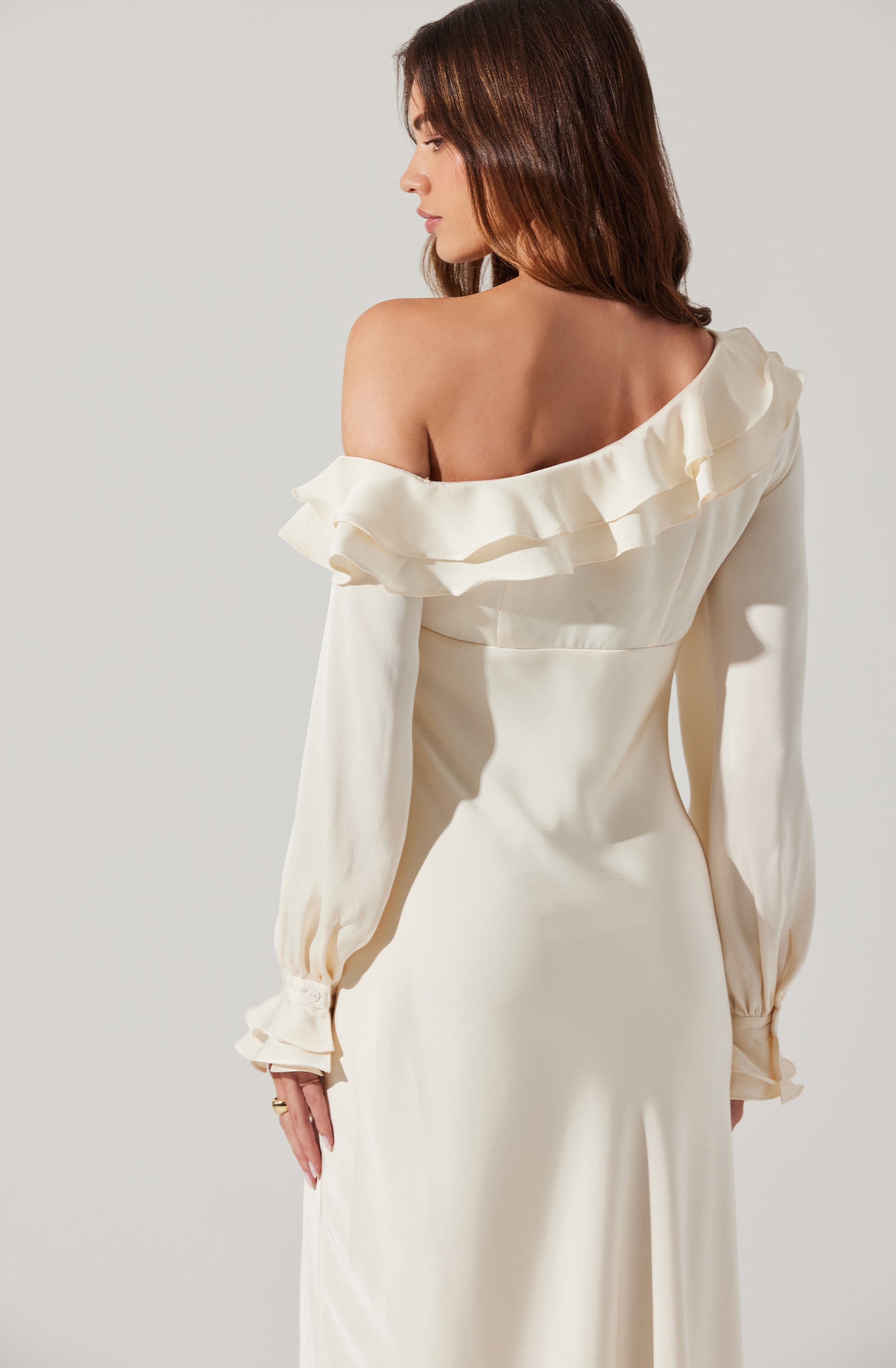 A woman is wearing an elegant off-the-shoulder cream-colored dress with ruffled detailing around the neckline. The dress has long, slightly puffed sleeves with gathered cuffs. The fabric appears smooth and satin-like, giving it a luxurious look.