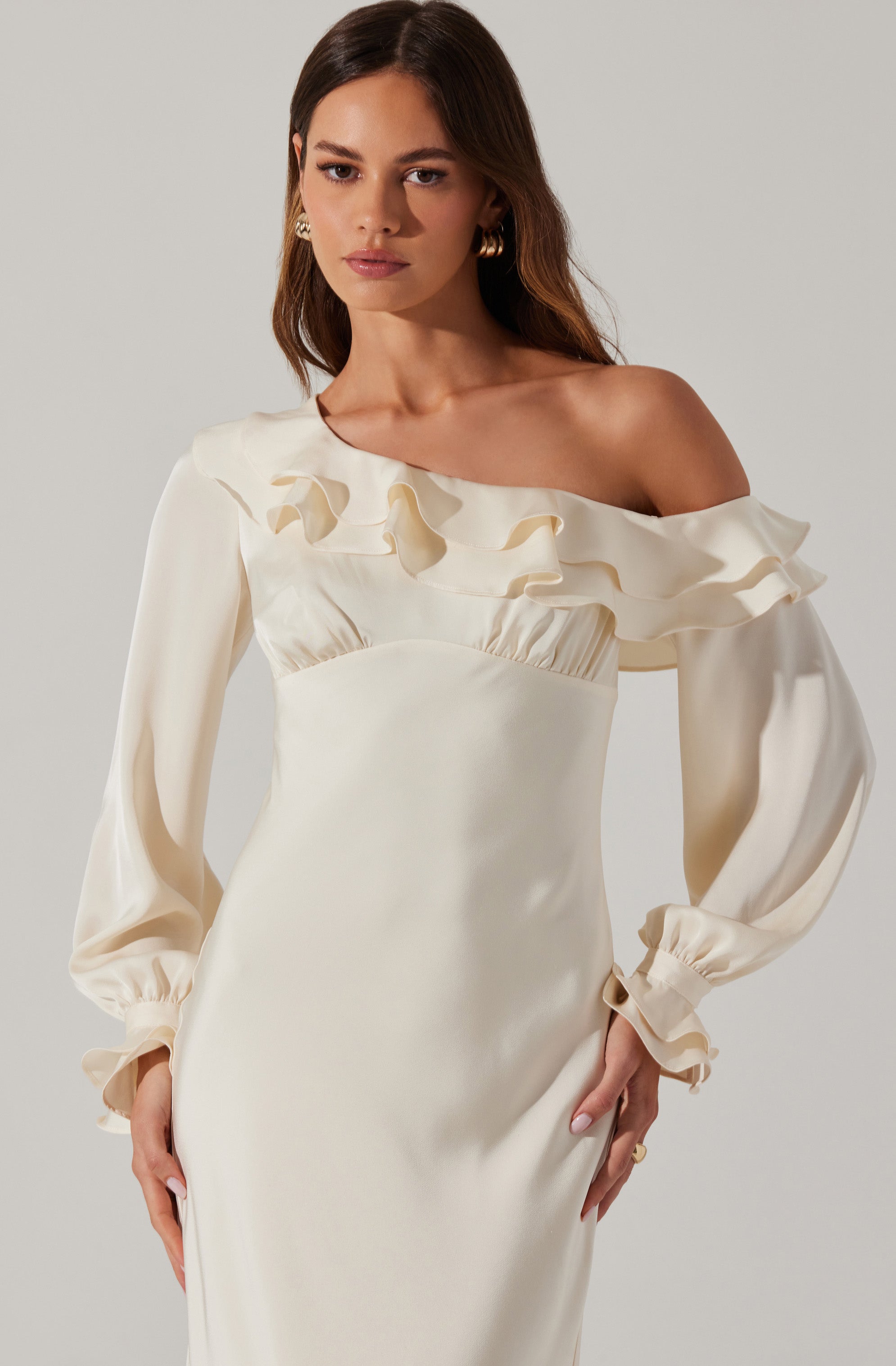 A woman is wearing an elegant, off-white, satin dress featuring a one-shoulder design with ruffled detailing along the neckline and shoulder. The dress has long, puffed sleeves with gathered cuffs.