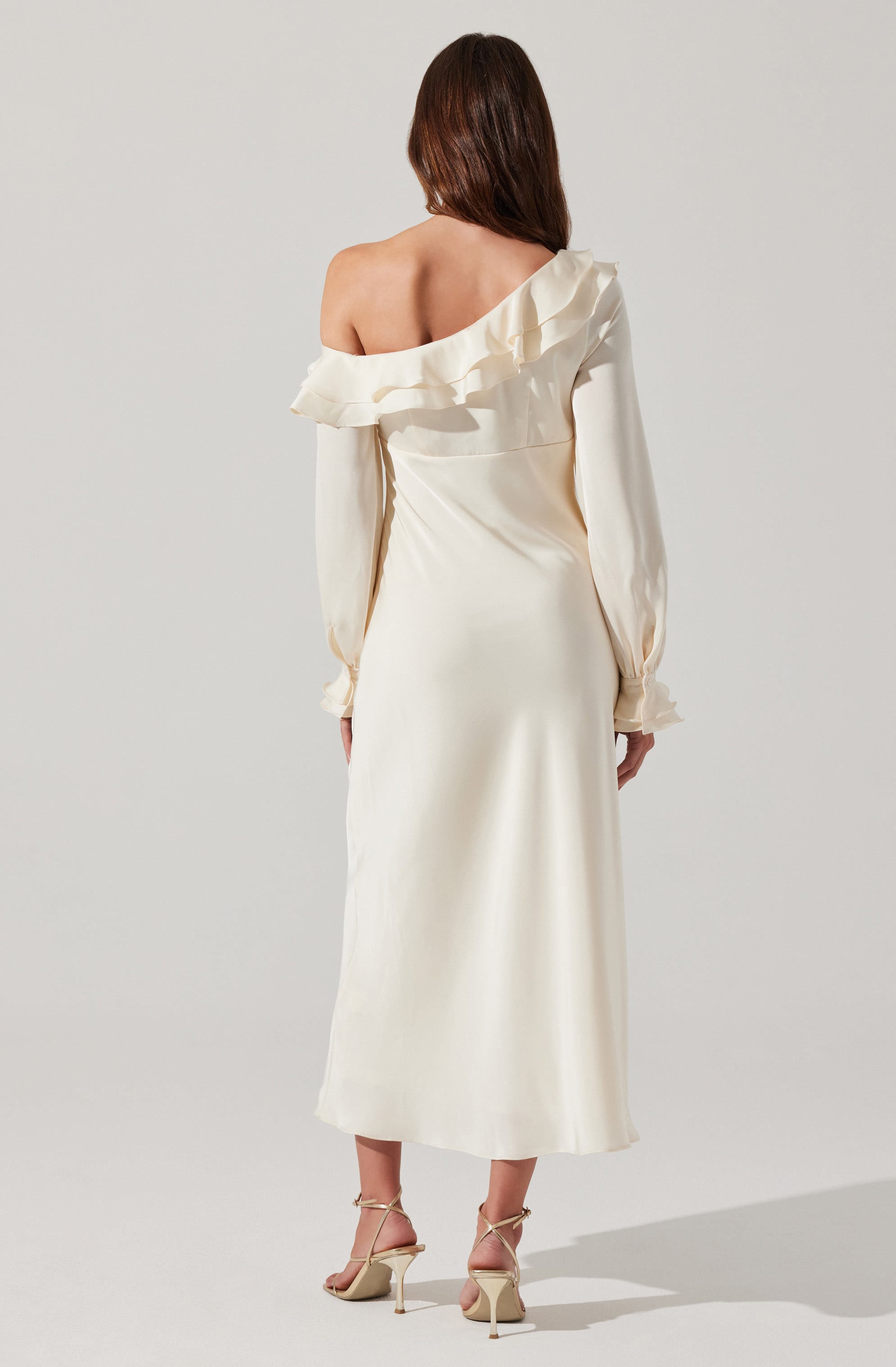 A woman is wearing an elegant, off-the-shoulder, cream-colored dress with long, billowy sleeves. The dress has a ruffled neckline that drapes over one shoulder, creating asymmetry. She is also wearing cream high-heeled sandals with ankle straps.