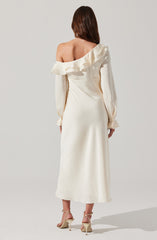 A woman is wearing an elegant, off-the-shoulder, cream-colored dress with long, billowy sleeves. The dress has a ruffled neckline that drapes over one shoulder, creating asymmetry. She is also wearing cream high-heeled sandals with ankle straps.