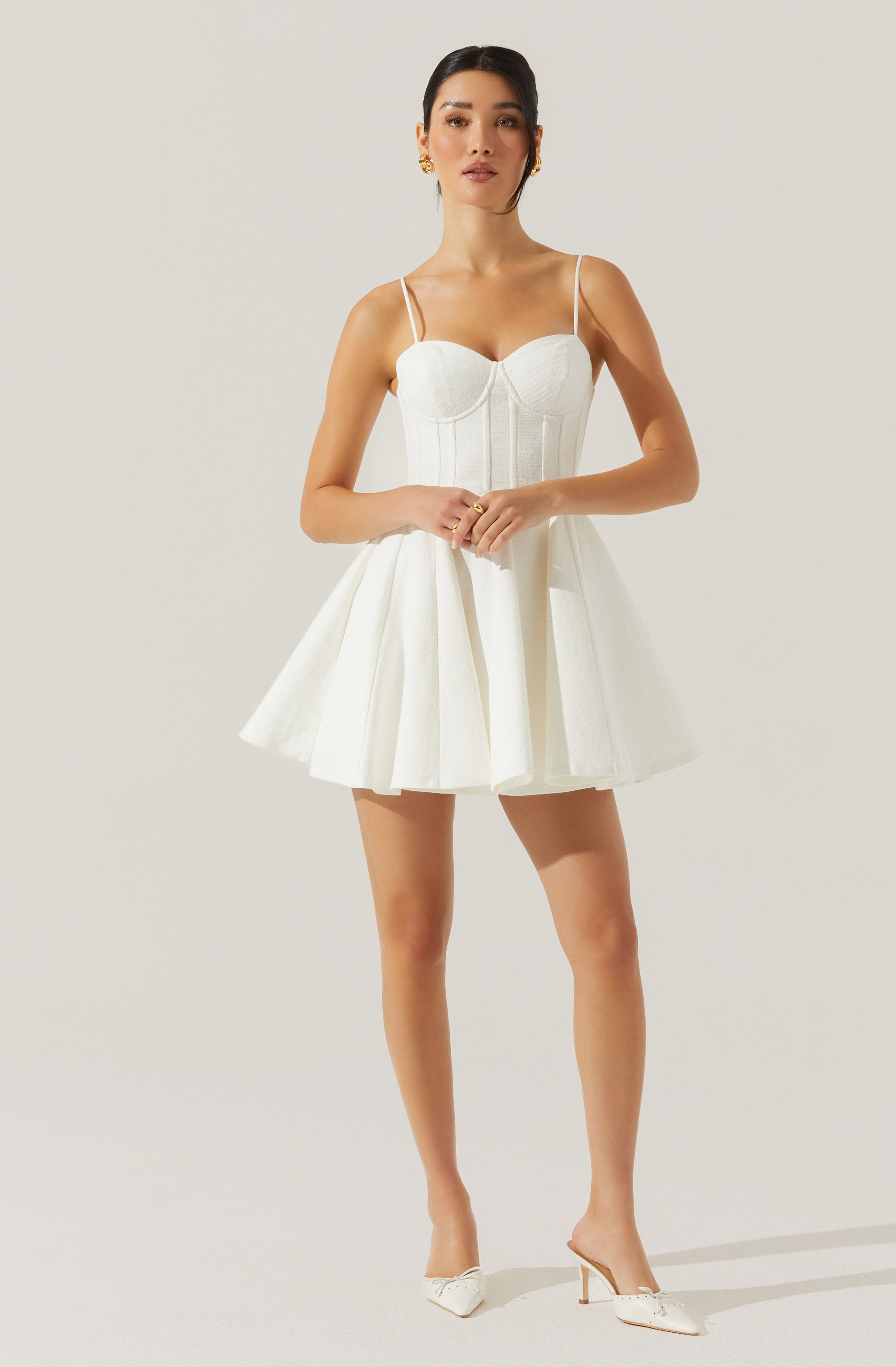A woman is wearing a white dress with a fitted bodice featuring vertical seams and a sweetheart neckline. The dress has thin spaghetti straps and a full, flared skirt with pleats. She is also wearing white high-heeled shoes with a decorative bow detail on the toe.