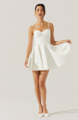 A woman is wearing a white dress with a fitted, bustier-style bodice featuring vertical seams and thin spaghetti straps. The dress has a full, pleated skirt that flares out from the waist and falls above the knees. She is also wearing white pointed-toe heels with decorative bows on the front.
