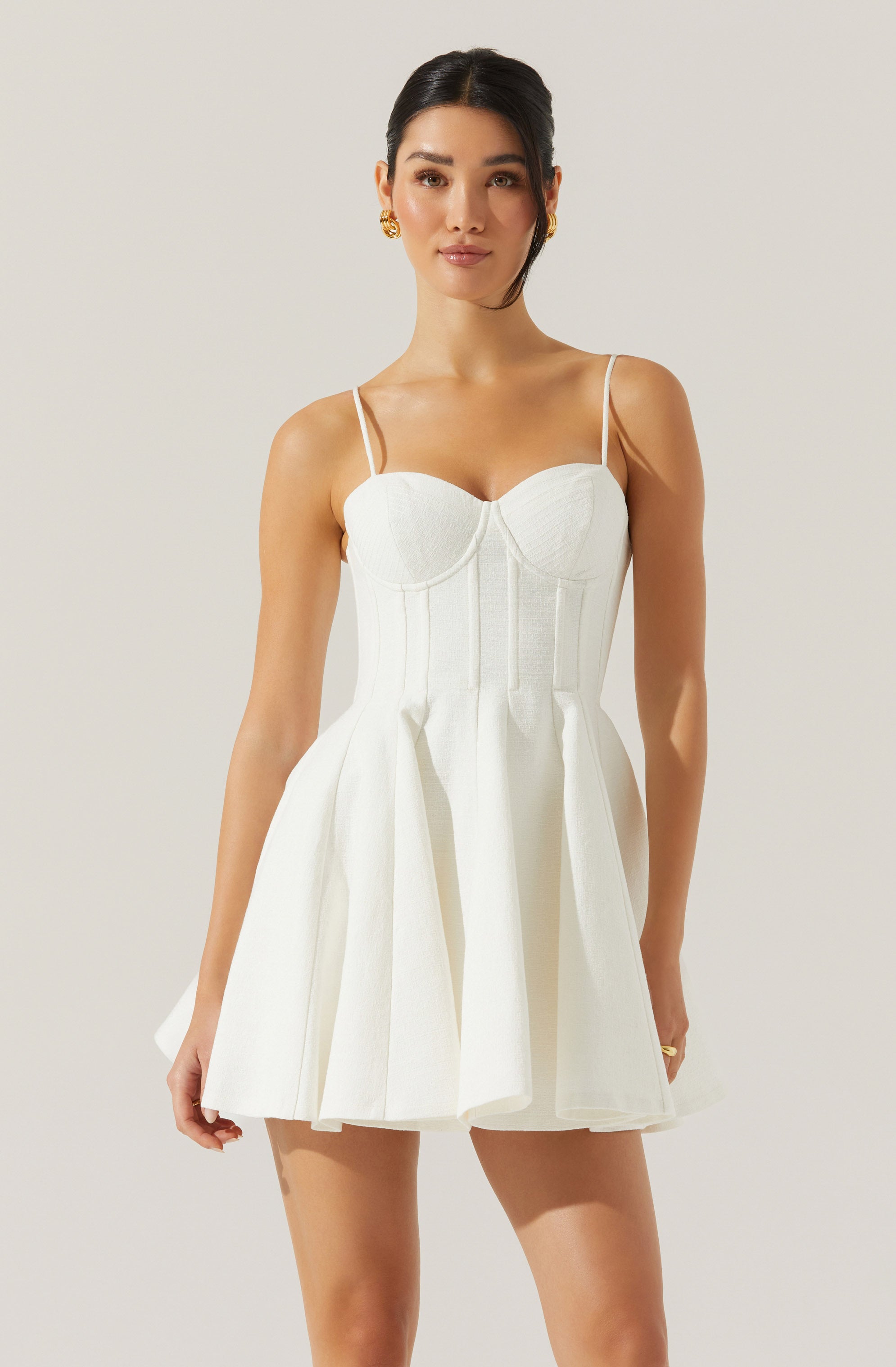 A woman is wearing a sleeveless, white dress with thin spaghetti straps. The dress features a fitted bodice with subtle vertical seam details and a flared, pleated skirt that reaches mid-thigh.