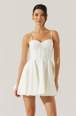 A woman is wearing a sleeveless, white dress with thin spaghetti straps. The dress features a fitted bodice with subtle vertical seam details and a flared, pleated skirt that reaches mid-thigh.
