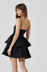 A young woman is wearing a black dress with a fitted bodice and thin spaghetti straps. The dress features a prominent, layered peplum at the waist and a short, flared skirt.