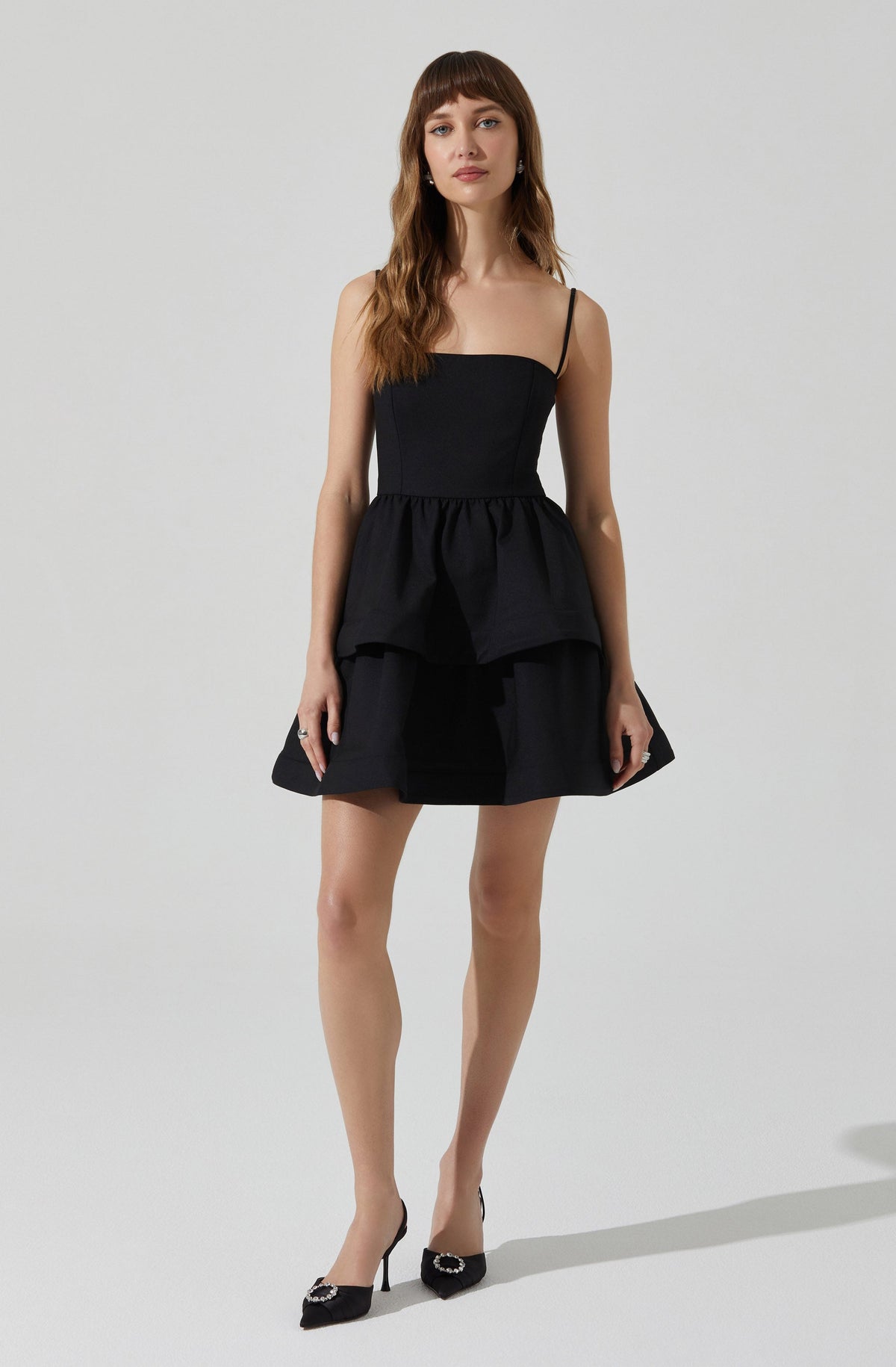 A woman is wearing a black dress with a fitted bodice and thin spaghetti straps. The dress features a peplum-style overlay at the waist and a flared skirt. She is also wearing black high-heeled shoes with decorative embellishments on the toes.