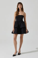 A woman is wearing a black dress with a fitted bodice and thin spaghetti straps. The dress features a peplum-style overlay at the waist and a flared skirt. She is also wearing black high-heeled shoes with decorative embellishments on the toes.