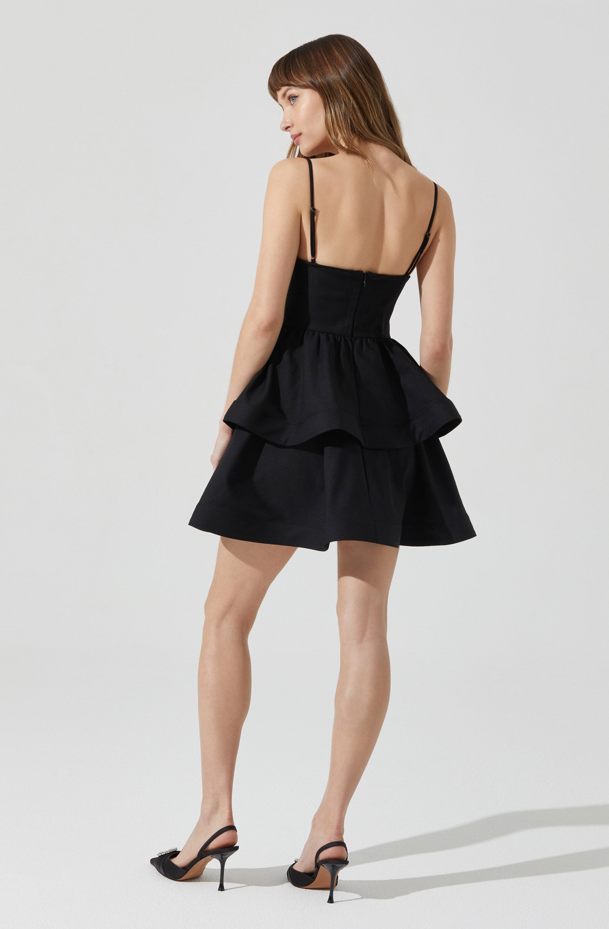 A woman is wearing a black sleeveless dress with thin spaghetti straps. The dress features a fitted bodice and a ruffled peplum waist that extends into a layered, flared skirt. She is also wearing black high-heeled shoes with ankle straps.