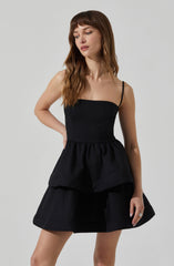 A woman is wearing a black sleeveless top with thin spaghetti straps and a peplum waist. She pairs it with a black skirt that has a layered, flared design.