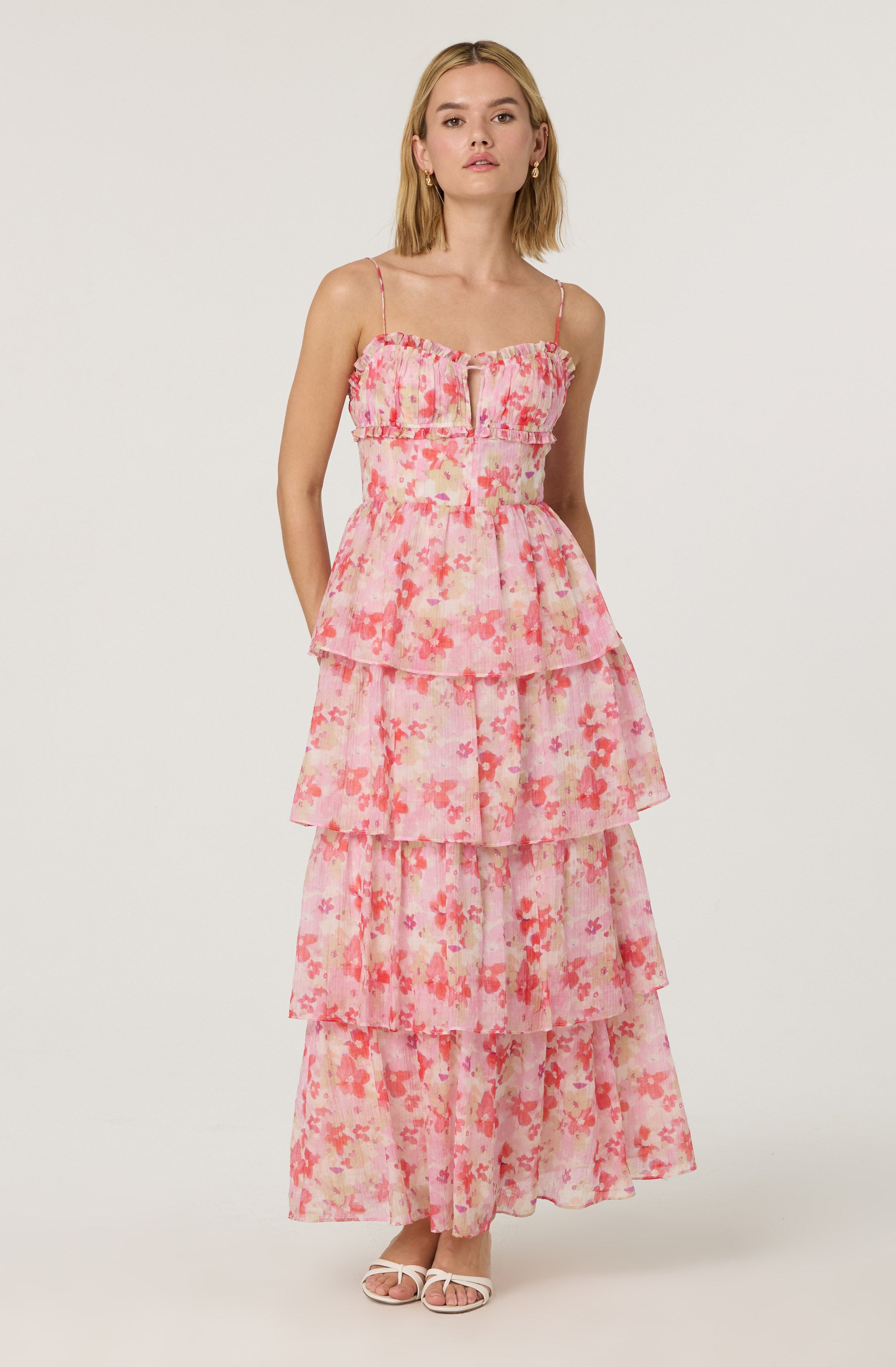 A woman is wearing a sleeveless, tiered maxi dress with thin spaghetti straps. The dress features a ruffled bodice with a zipper in the front, and it has a pink floral pattern with shades of pink, peach, and coral on a sheer fabric. The dress has multiple layered sections, creating a flowing, bohemian look. She is also wearing light-colored open-toe sandals.