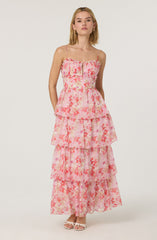 A woman is wearing a sleeveless, tiered maxi dress with thin spaghetti straps. The dress features a ruffled bodice with a zipper in the front, and it has a pink floral pattern with shades of pink, peach, and coral on a sheer fabric. The dress has multiple layered sections, creating a flowing, bohemian look. She is also wearing light-colored open-toe sandals.
