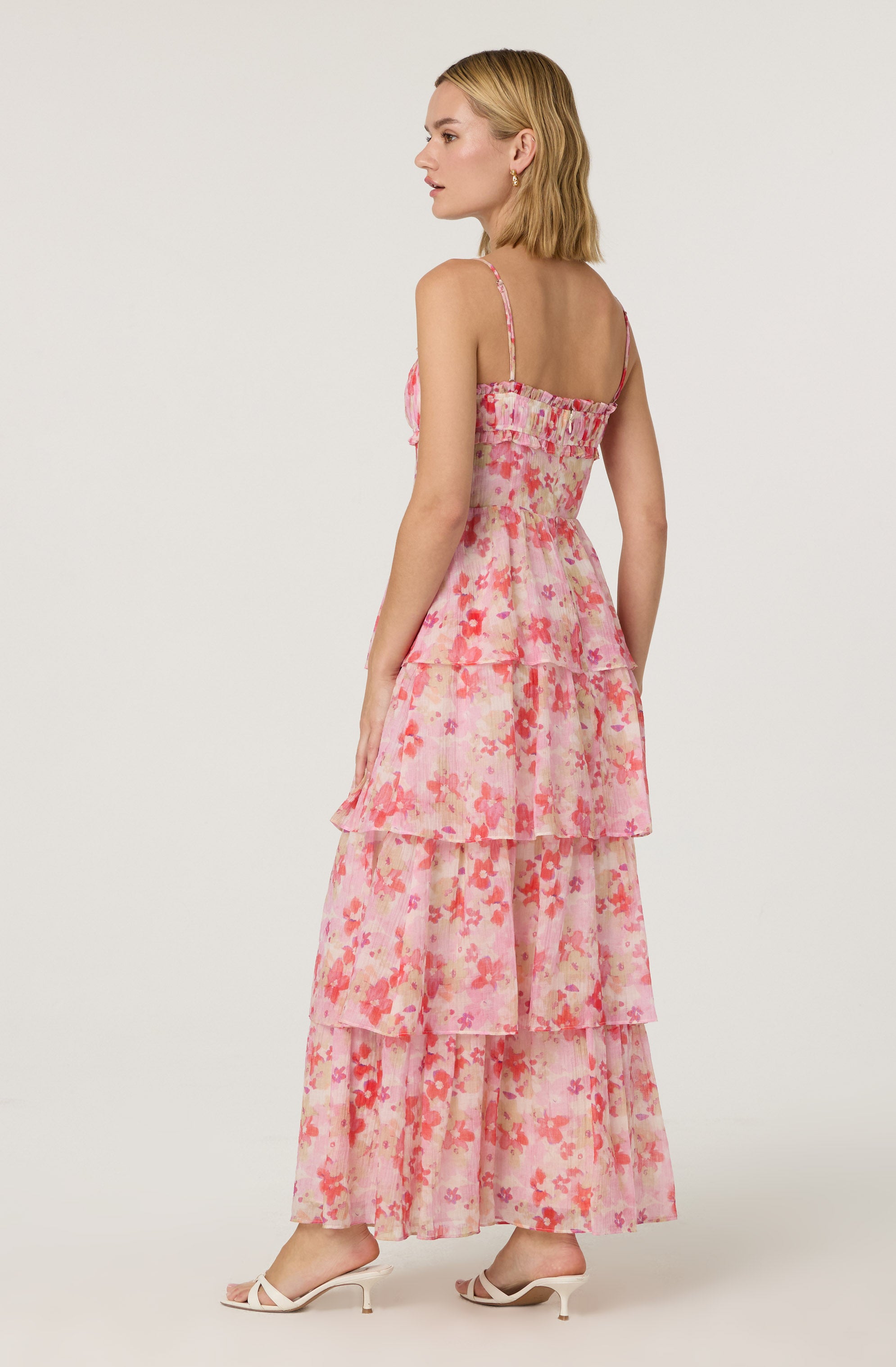 A woman is wearing a sleeveless, ankle-length dress with thin spaghetti straps. The dress features a pink and coral floral pattern with multiple tiered ruffles. She is also wearing white, open-toe high-heeled sandals.