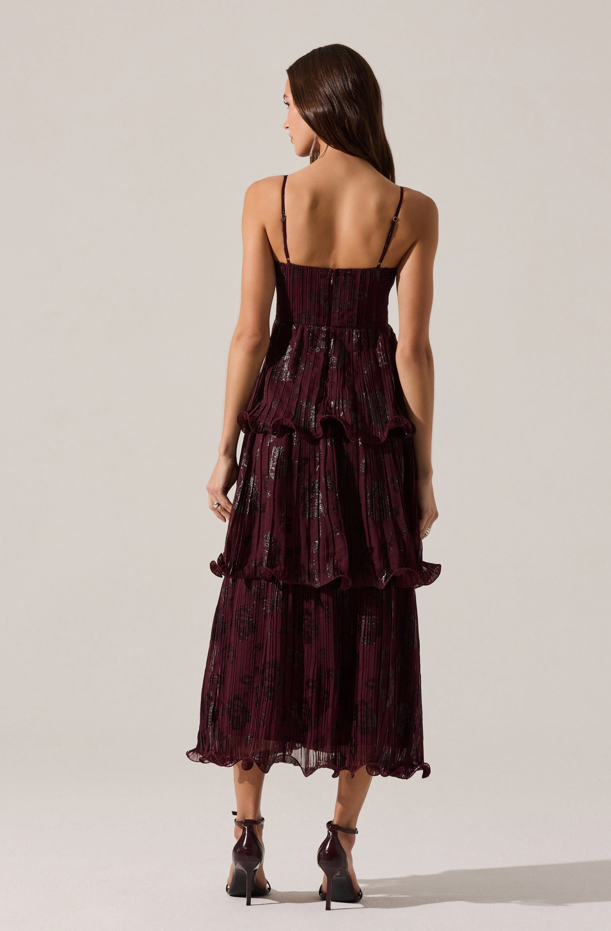 A woman is wearing a sleeveless, full-length dress with thin spaghetti straps. The dress is deep burgundy with a shiny, textured pattern, featuring multiple layered, ruffled tiers that add volume and movement. The fabric appears to be lightweight and semi-transparent with a delicate, slightly crinkled appearance. She pairs the dress with dark high-heeled shoes with ankle straps.