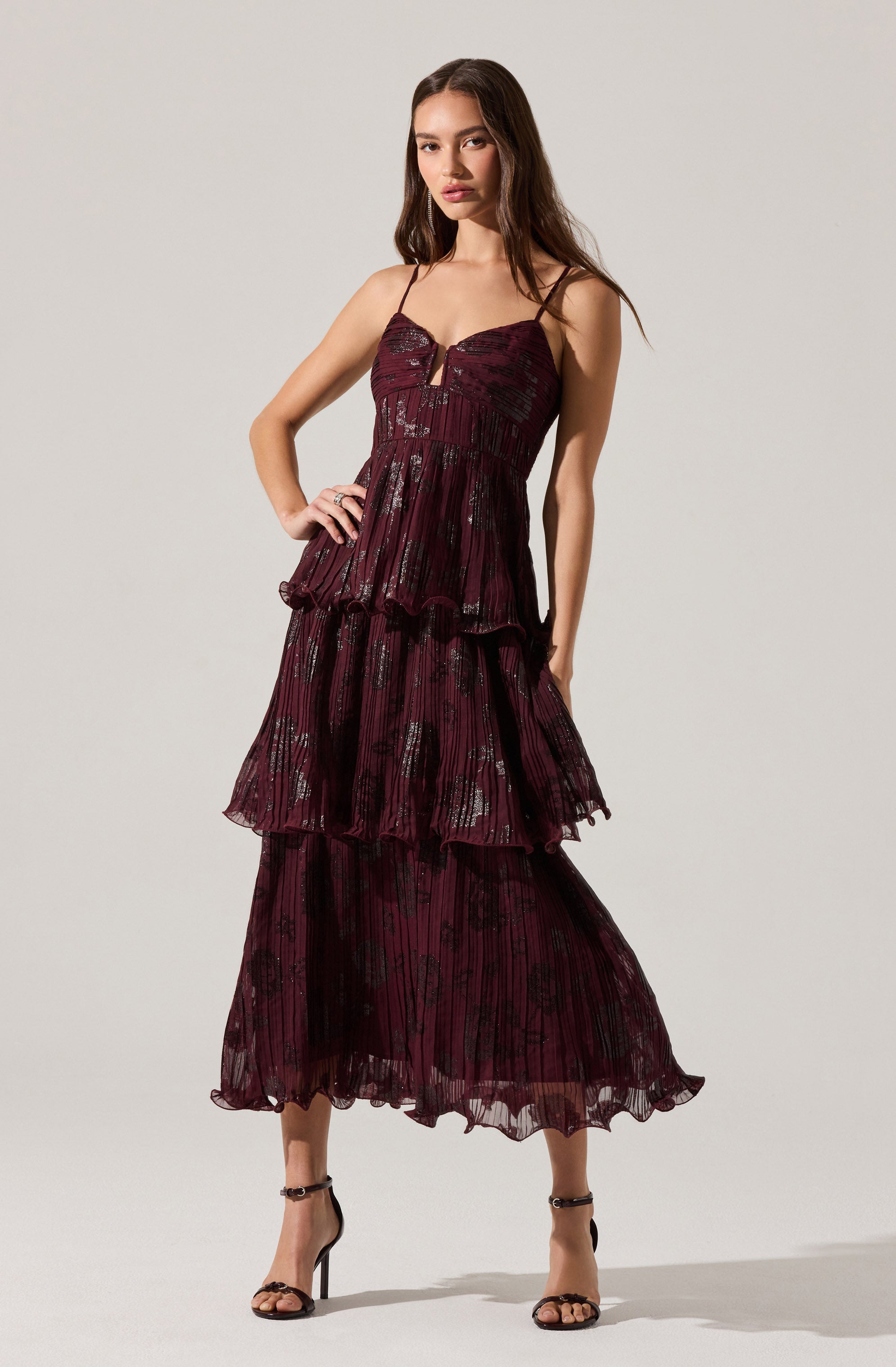 A woman is wearing a deep burgundy-colored dress with thin spaghetti straps. The dress features a fitted bodice with a sweetheart neckline and a small vertical slit at the center. The skirt has multiple layered, ruffled tiers made from a sheer, lightweight fabric with a subtle floral and striped pattern in black. She is also wearing black high-heeled sandals with ankle straps.