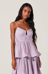 A woman is wearing a sleeveless, lavender-colored dress with thin spaghetti straps. The dress features a pleated fabric with vertical stripes, a fitted bodice with a small keyhole opening at the center, and a peplum-style waist that flares out over the matching pleated skirt.