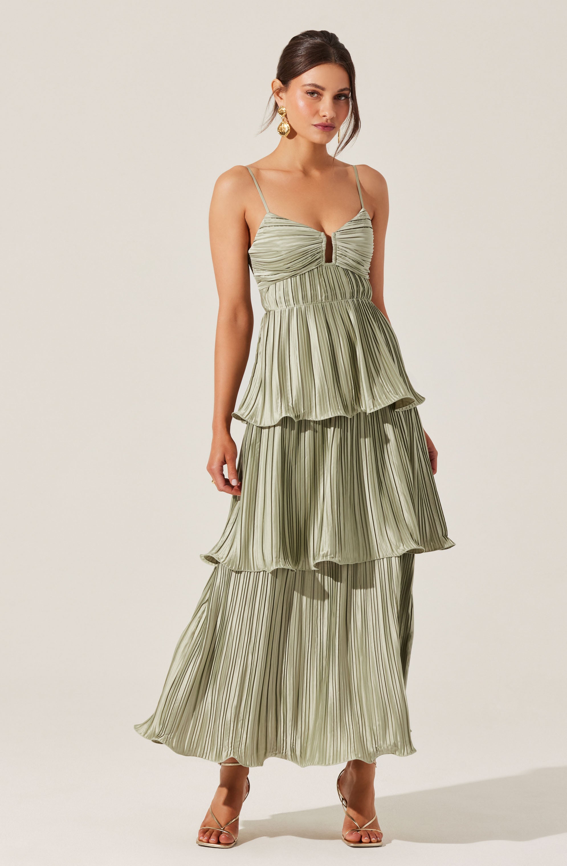 A woman is wearing a light green, pleated maxi dress with thin spaghetti straps. The dress features a ruched bodice with a small keyhole cutout in the center. The skirt has three tiers, each with a ruffled edge, creating a flowing, layered look. She is accessorized with large gold earrings and nude, strappy high-heeled sandals.