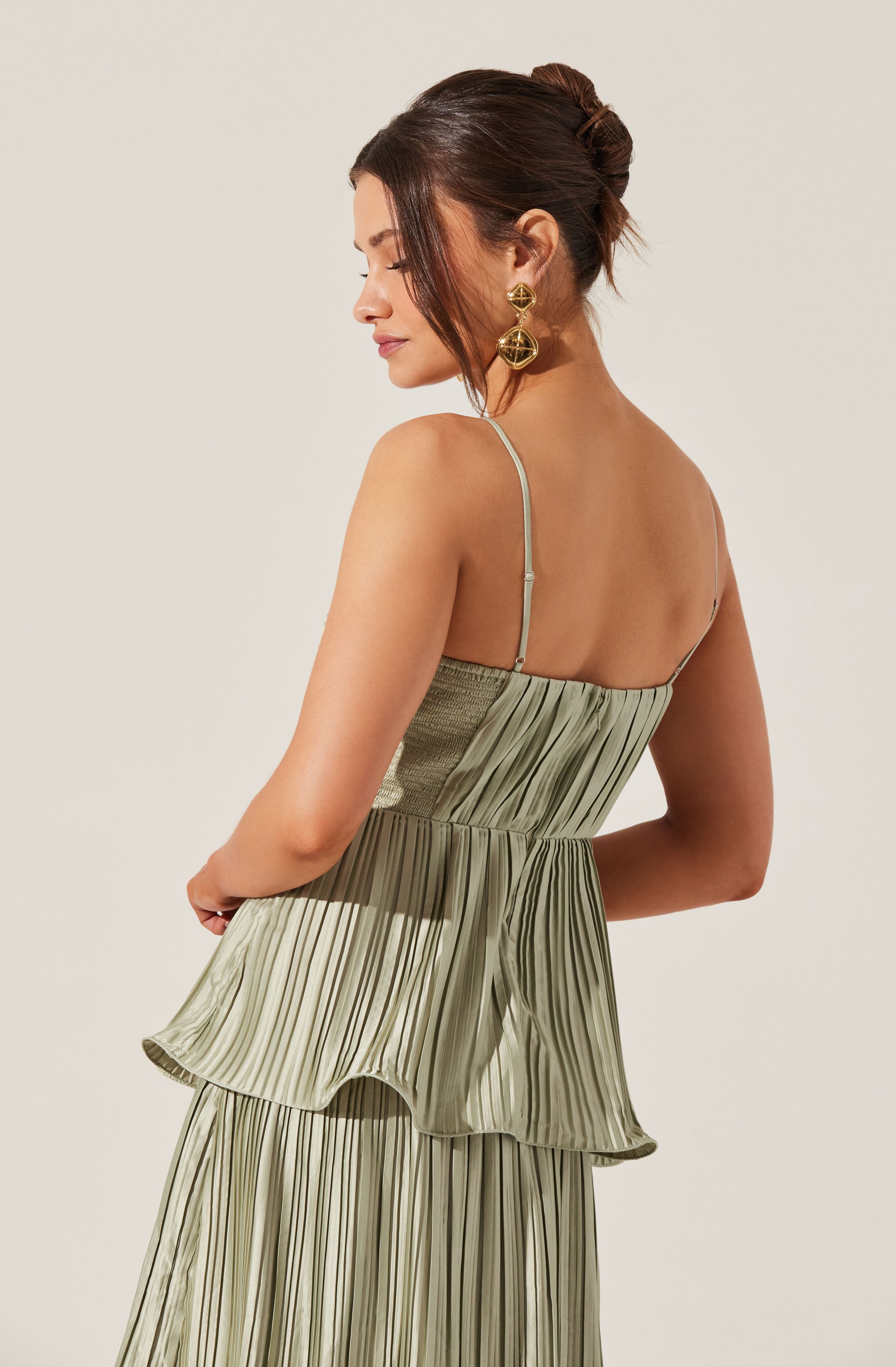 A woman is wearing a light green, pleated, sleeveless dress with thin spaghetti straps. The dress has a layered, ruffled overlay on the skirt portion. She is accessorized with large, statement gold earrings and has her dark hair styled in an elegant updo.
