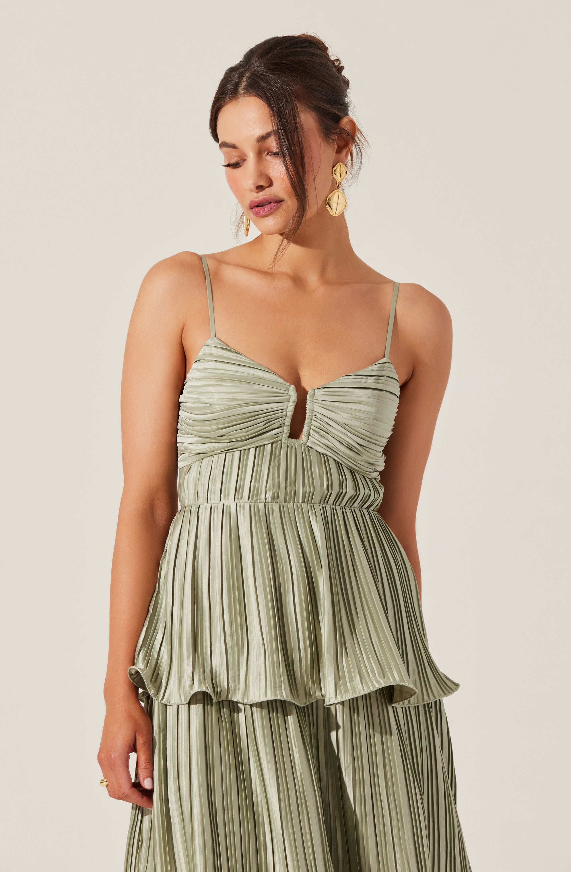 A woman is wearing a light green, satin-like, pleated dress with thin spaghetti straps. The top has a ruched bust with a small keyhole opening in the center, and the skirt features layered tiers with vertical stripes. She has short, dark hair styled in a loose updo, and is accessorized with large, gold, geometric earrings.