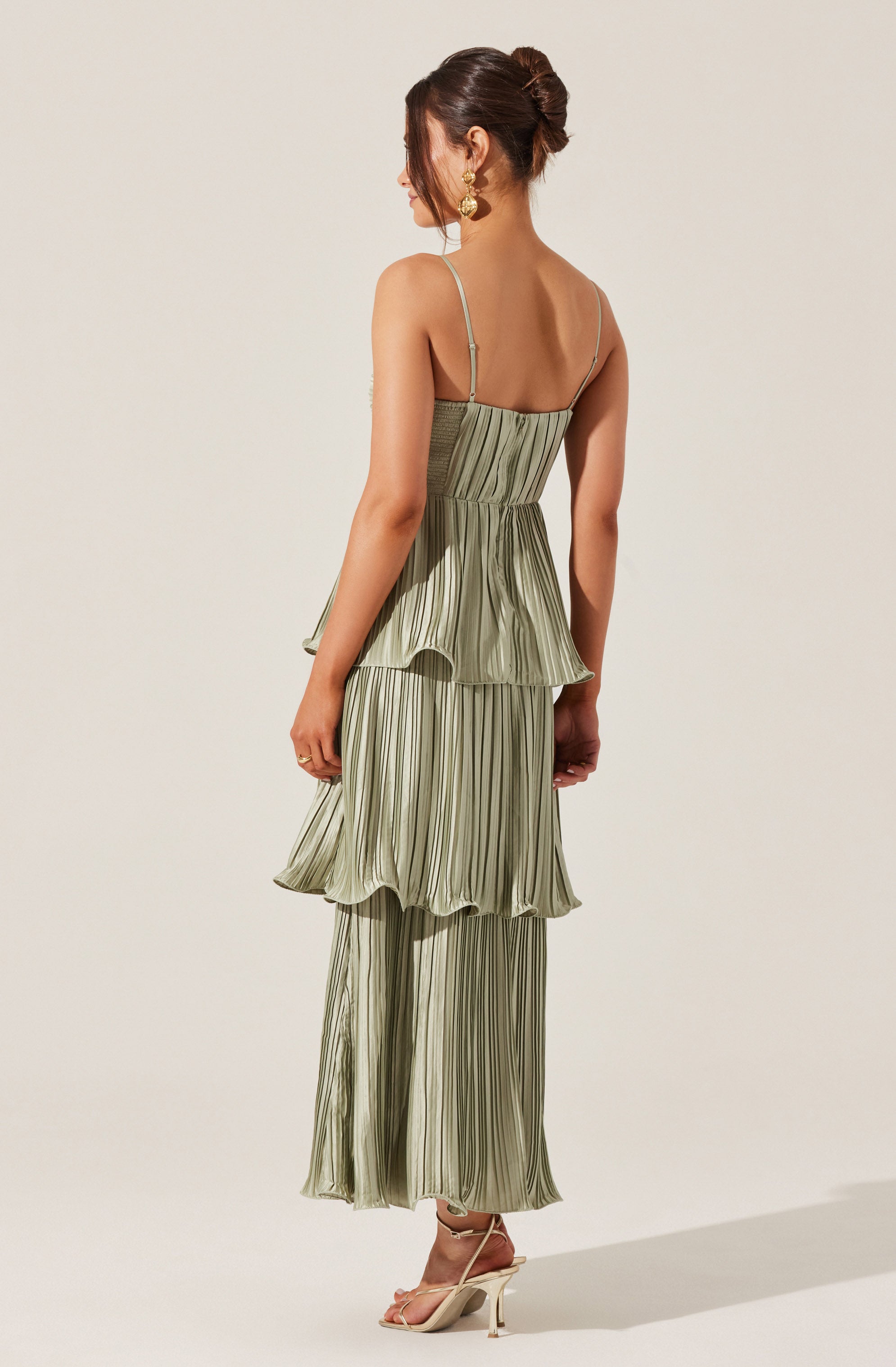 A woman is wearing a light green, pleated, strap-shoulder dress with thin spaghetti straps. The dress features a fitted bodice and a layered, ruffled skirt with vertical stripes. She has paired the dress with nude high-heeled sandals and gold earrings.