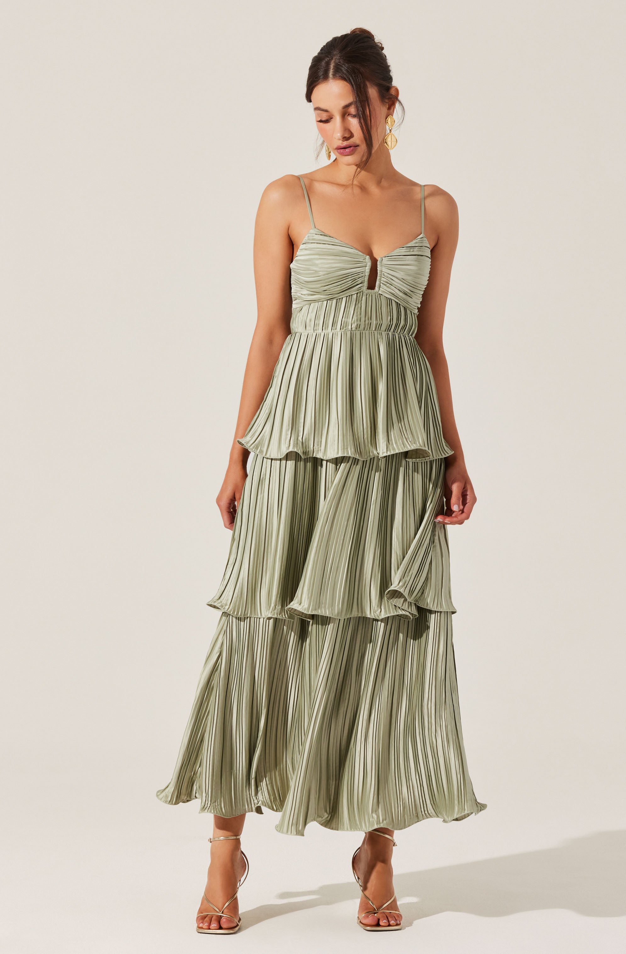 The woman is wearing a light green dress with vertical pleats and tiered layers. The dress has a fitted bodice with a slit in the front and thin spaghetti straps. She is also wearing nude high-heeled sandals and gold hoop earrings.