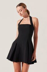 A woman is wearing a sleeveless black dress with a fitted bodice and a flared, pleated skirt. The dress has wide shoulder straps and a square neckline. She is accessorizing with gold statement earrings.