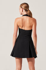 A woman is wearing a strapless black dress with a halter neck. The dress is fitted at the top and flares out into a pleated skirt. The dress has a zipper closure at the back. The woman has light brown hair styled in a low ponytail.
