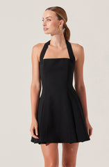 A woman is wearing a sleeveless black dress with a fitted bodice and an A-line skirt that falls above the knees. The dress features wide straps that go over her shoulders and connect to the back. She accessorizes with gold earrings.