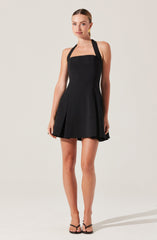 A woman is wearing a sleeveless black dress with wide shoulder straps and a square neckline. The dress has a fitted bodice and a flared skirt with pleats. She is also wearing black high-heeled sandals.