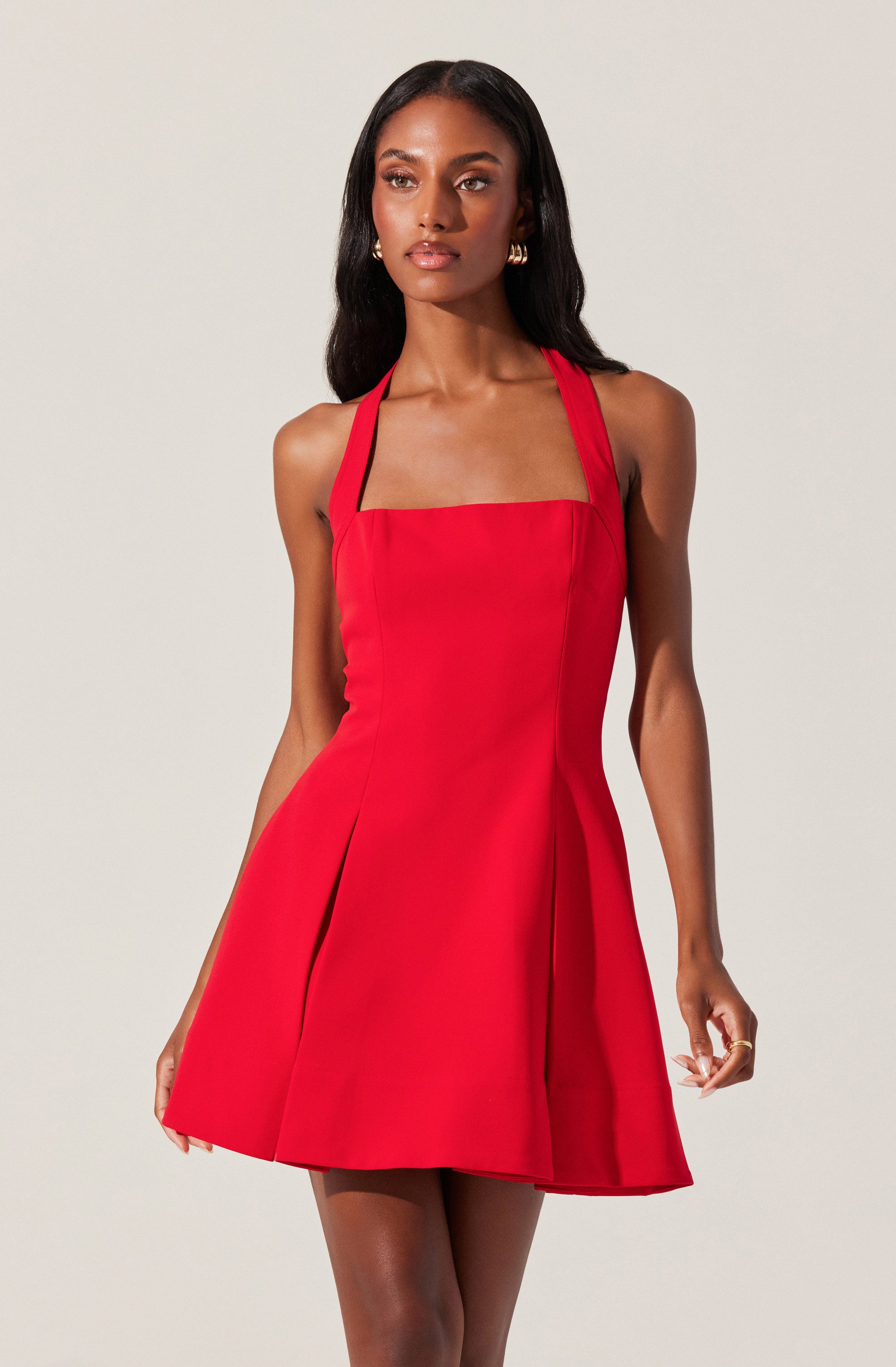 A woman is wearing a sleeveless, red dress with wide shoulder straps, a fitted bodice, and a flared, pleated skirt. The dress has a square neckline and structured seams along the front.