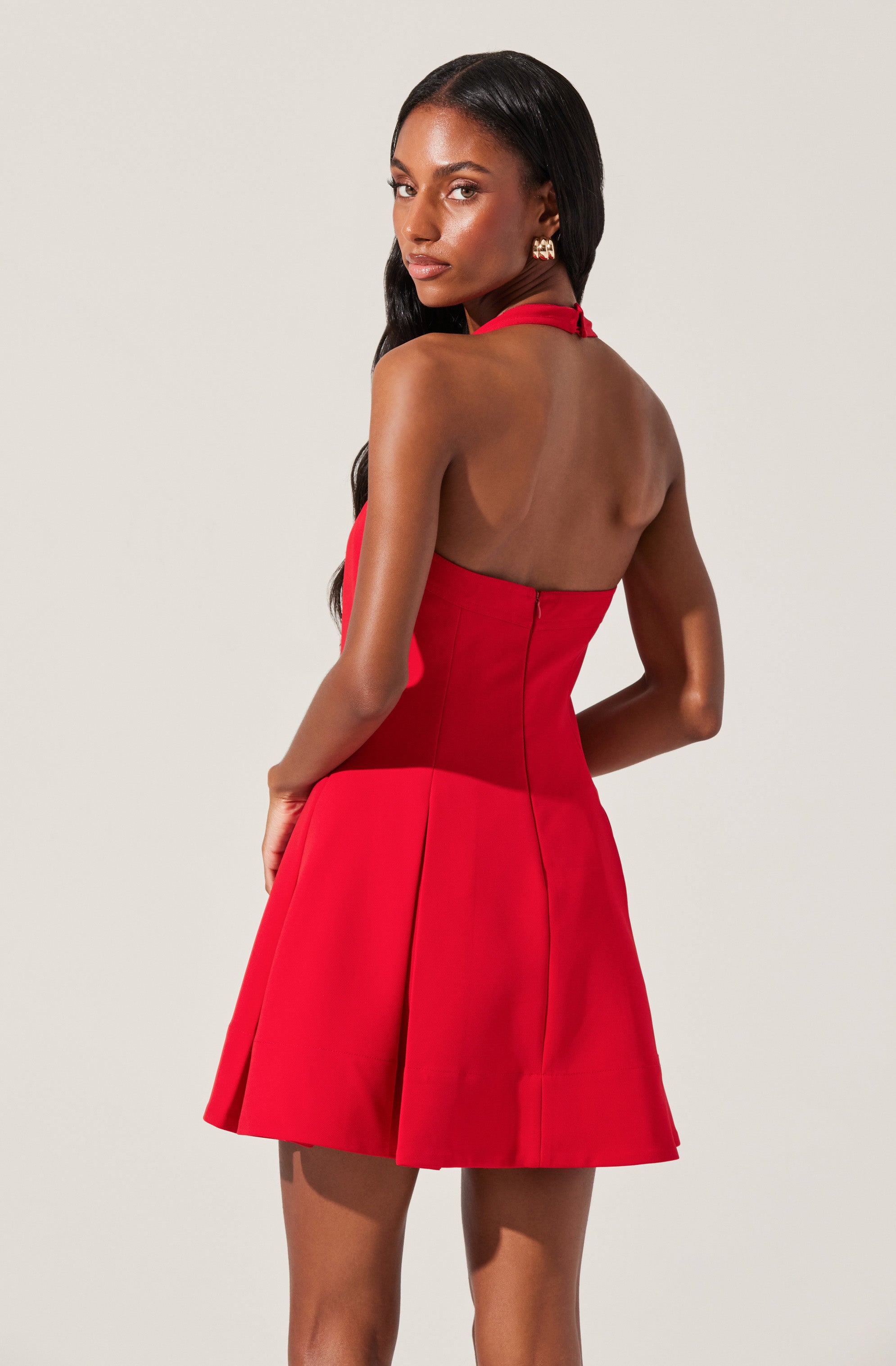 A woman is wearing a sleeveless, red dress with a halter neckline that ties around her neck. The dress is fitted at the waist and has a flared, pleated skirt. The back of the dress is open, exposing her shoulders and upper back.